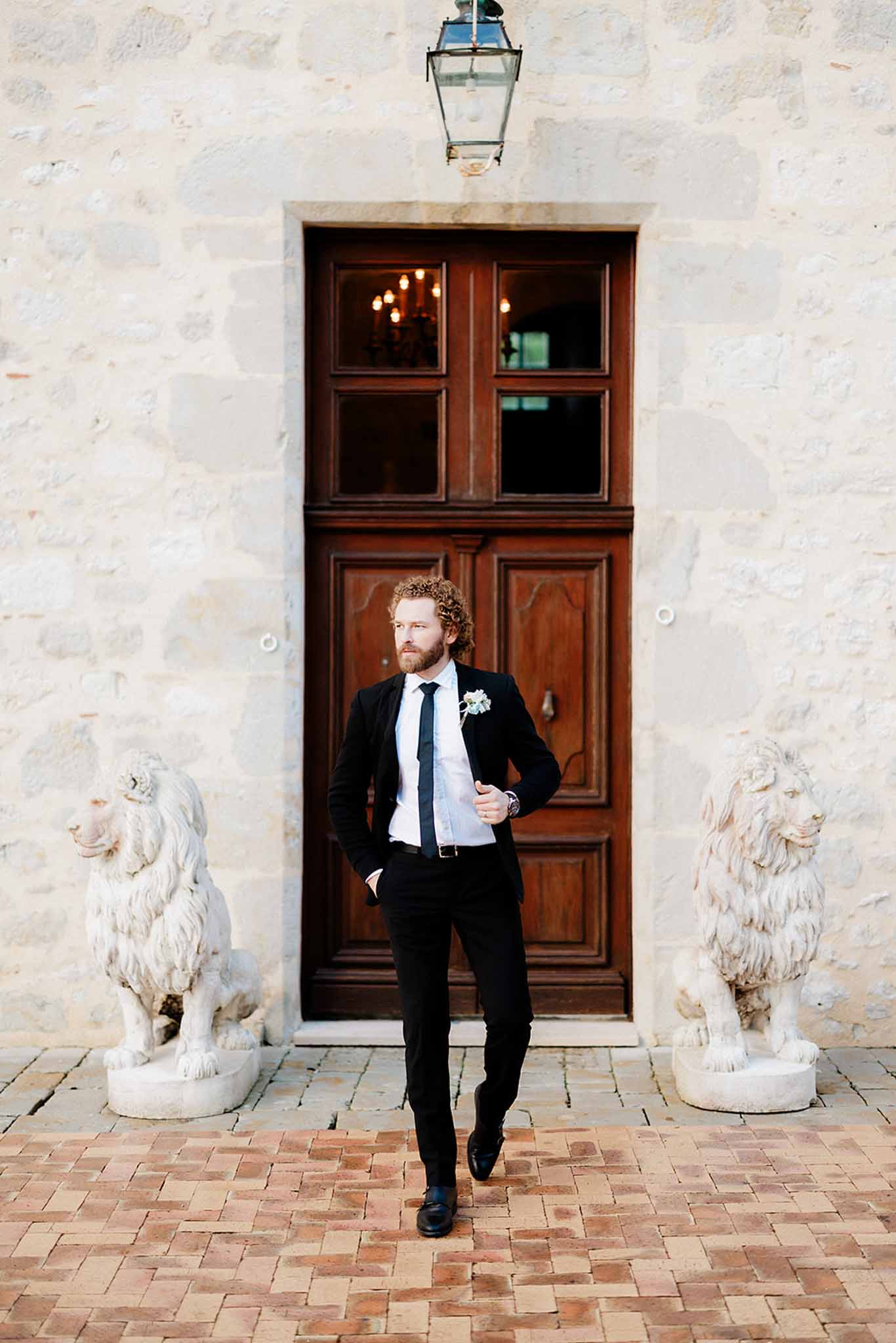 Groom in black suit posing between stone lion statues at chateau entrance with brass lantern