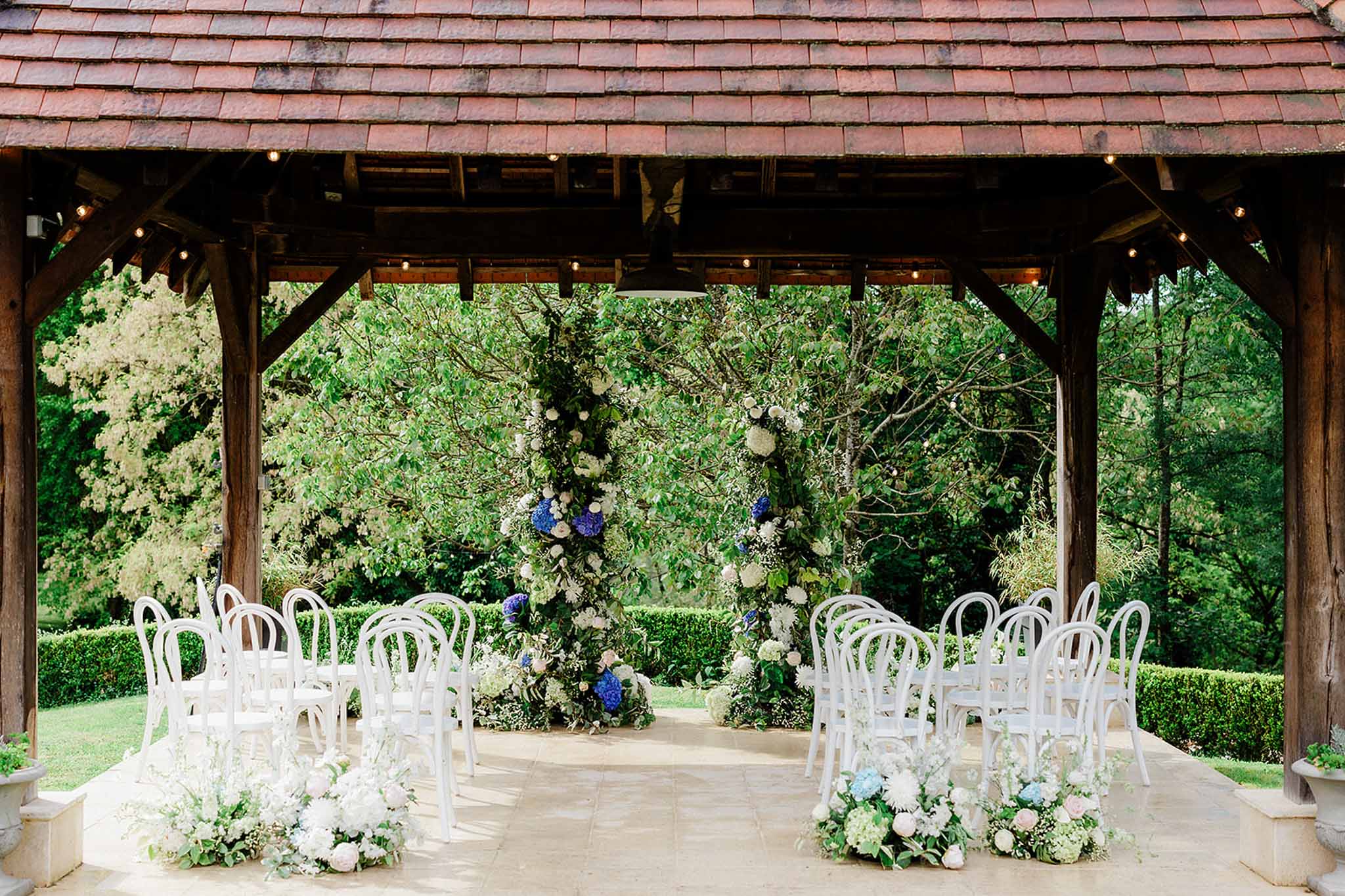 Ceremony under timber-frame pavilion, white Chiavari chairs, blue delphinium and white hydrangea backdrop, string lights
