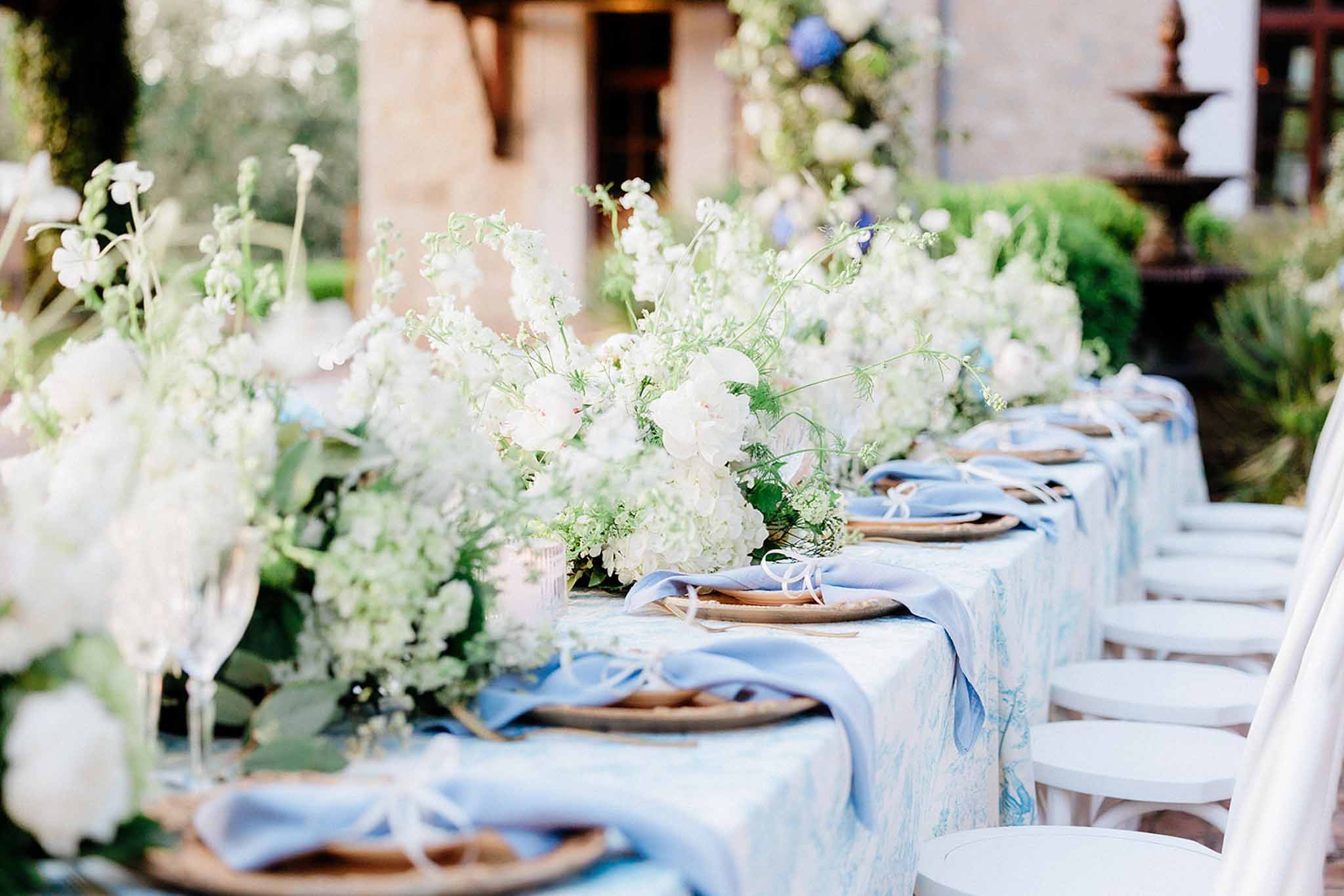 Long reception table with white linen, white flower centerpieces and blue napkins in a Mediterranean courtyard with stone buildings.