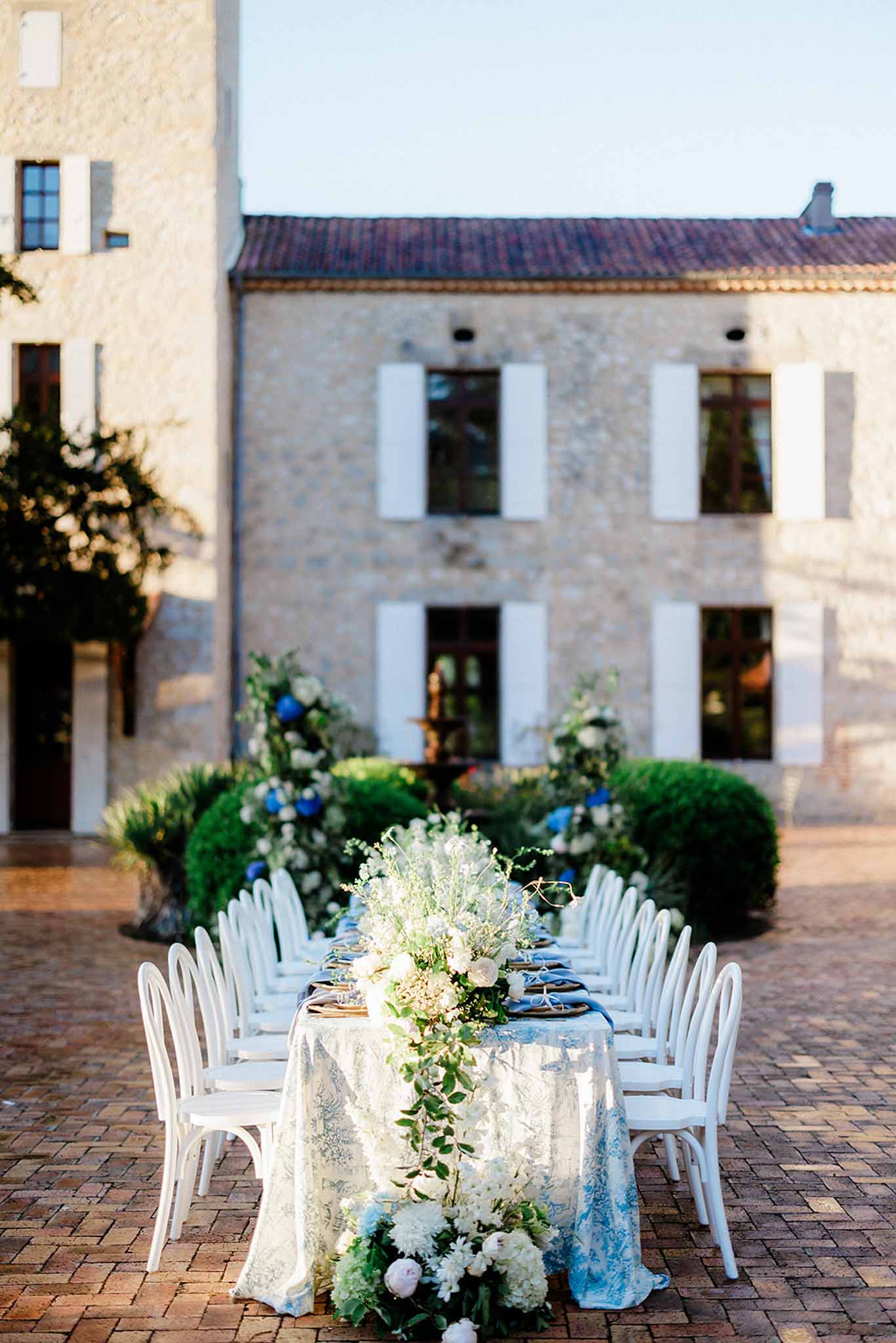 Outdoor reception table with lace overlay, blue florals, and cascading flower runner by stone building