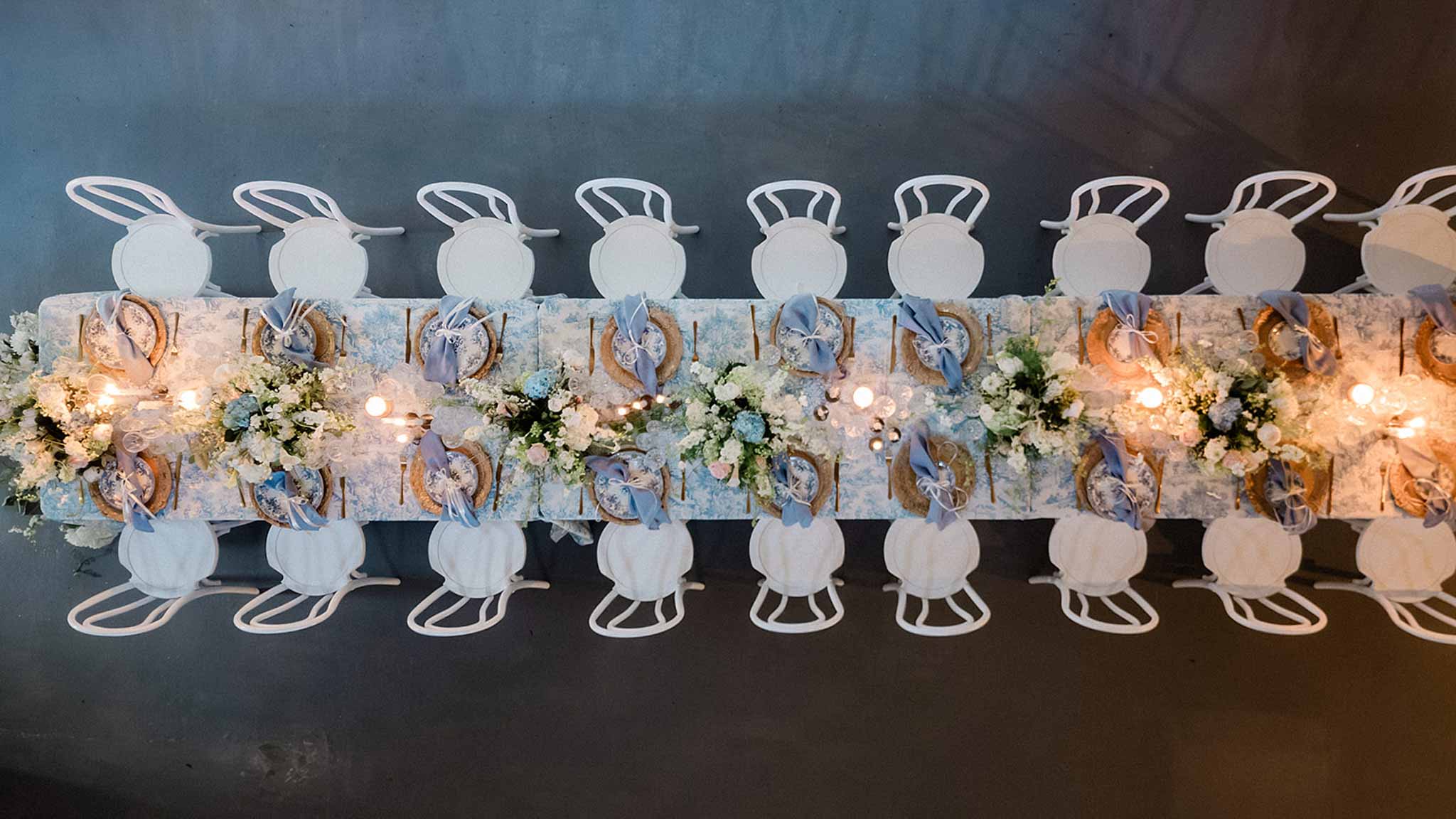 Aerial view of long reception banquet table with blue and gold runner, woven chargers, white florals and string lights