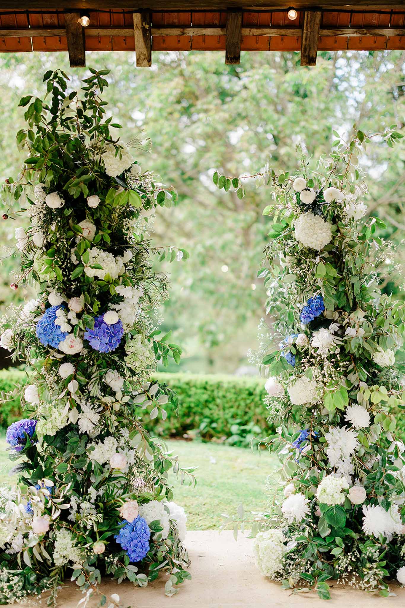 Two tall floral columns of white and blue hydrangeas flanking wooden pergola ceremony backdrop