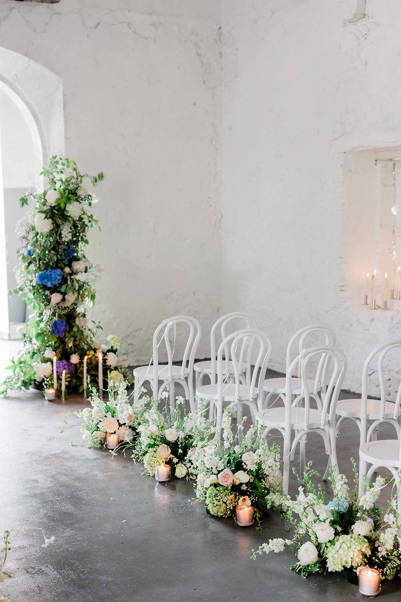 Ceremony setup in industrial white brick space with cream and blue hydrangea arch and aisle lined with white florals