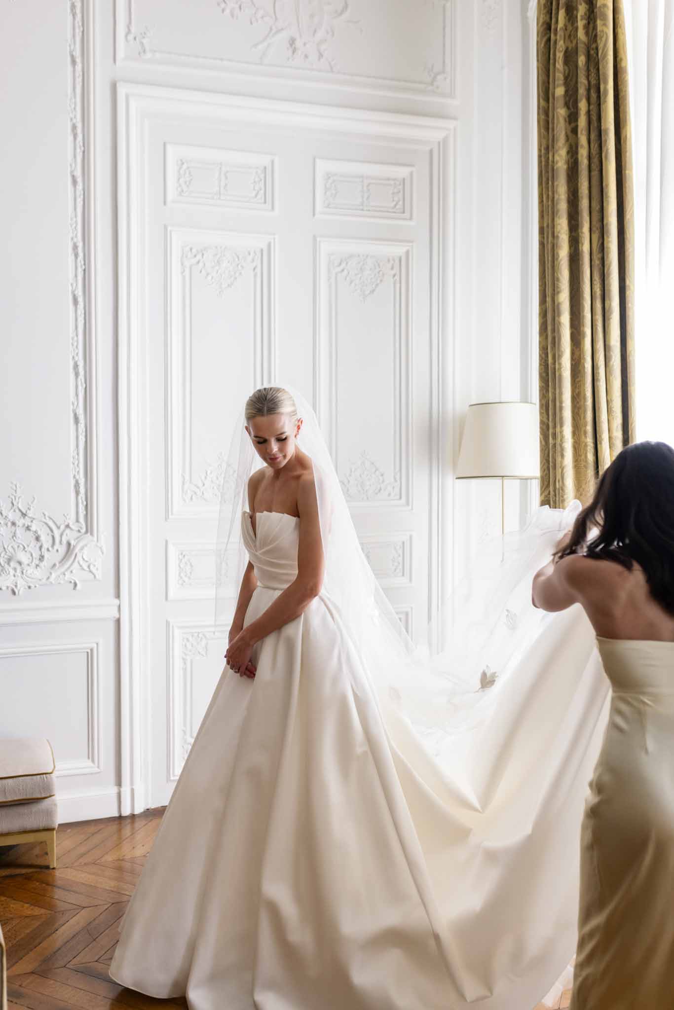 Bride in ivory strapless gown with attendant arranging train in neoclassical dressing room