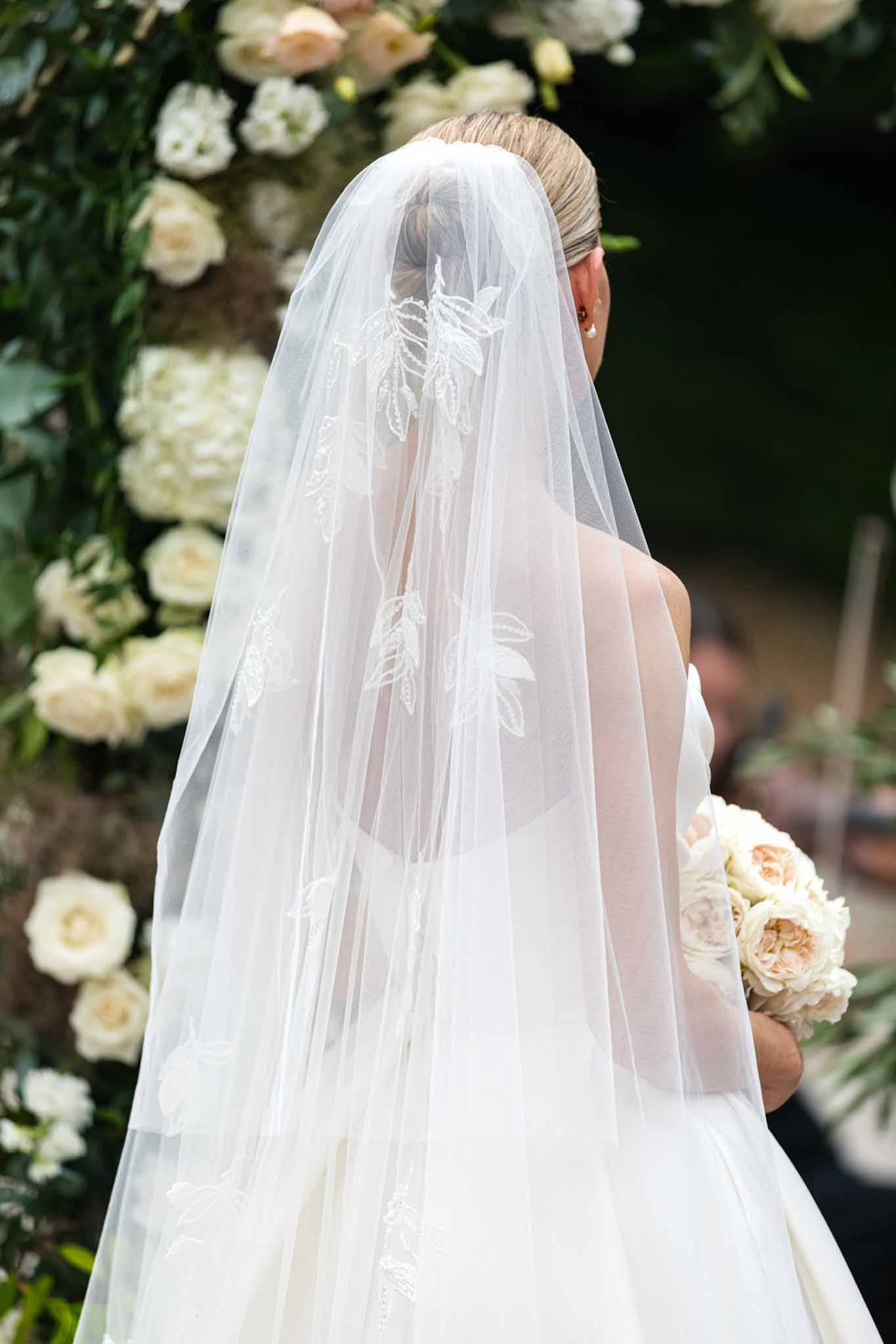 Bride seen from behind at ceremony wearing ivory embroidered veil, holding bouquet of ivory and peach roses