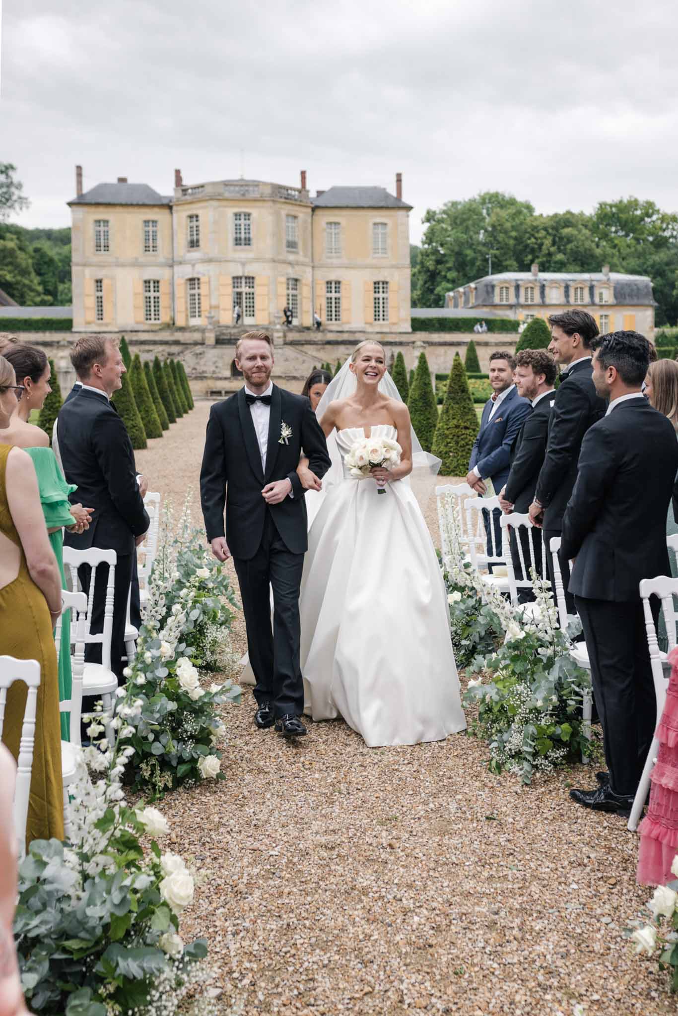 Bride and groom walking recessional in formal French chateau courtyard lined with white chairs and standing guests