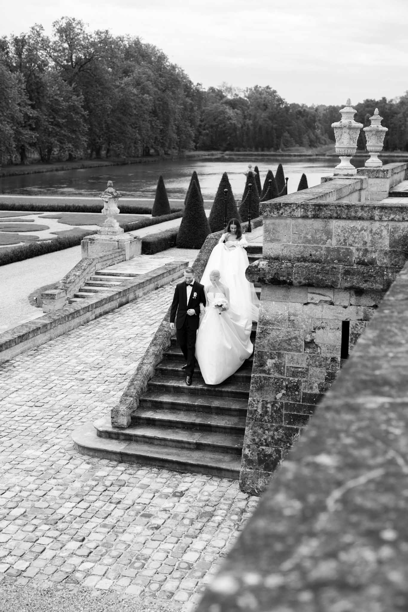 Black and white portrait of bride and groom descending formal garden staircase, dramatic train on steps, cone topiaries