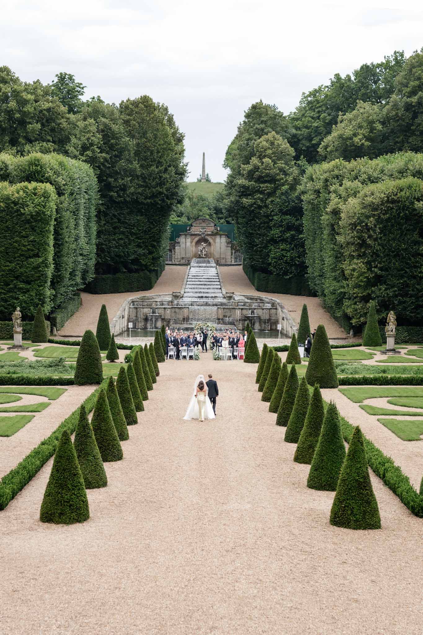 Bride and groom walk down formal gravel path lined with cone topiaries toward outdoor reception with fountain beyond