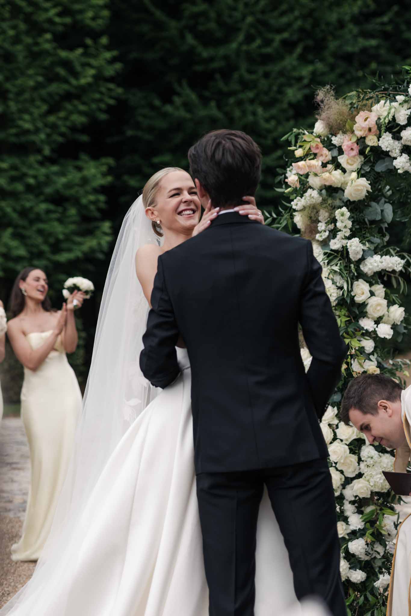 Bride and groom embracing after ceremony beside a tall installation of ivory roses and white hydrangeas