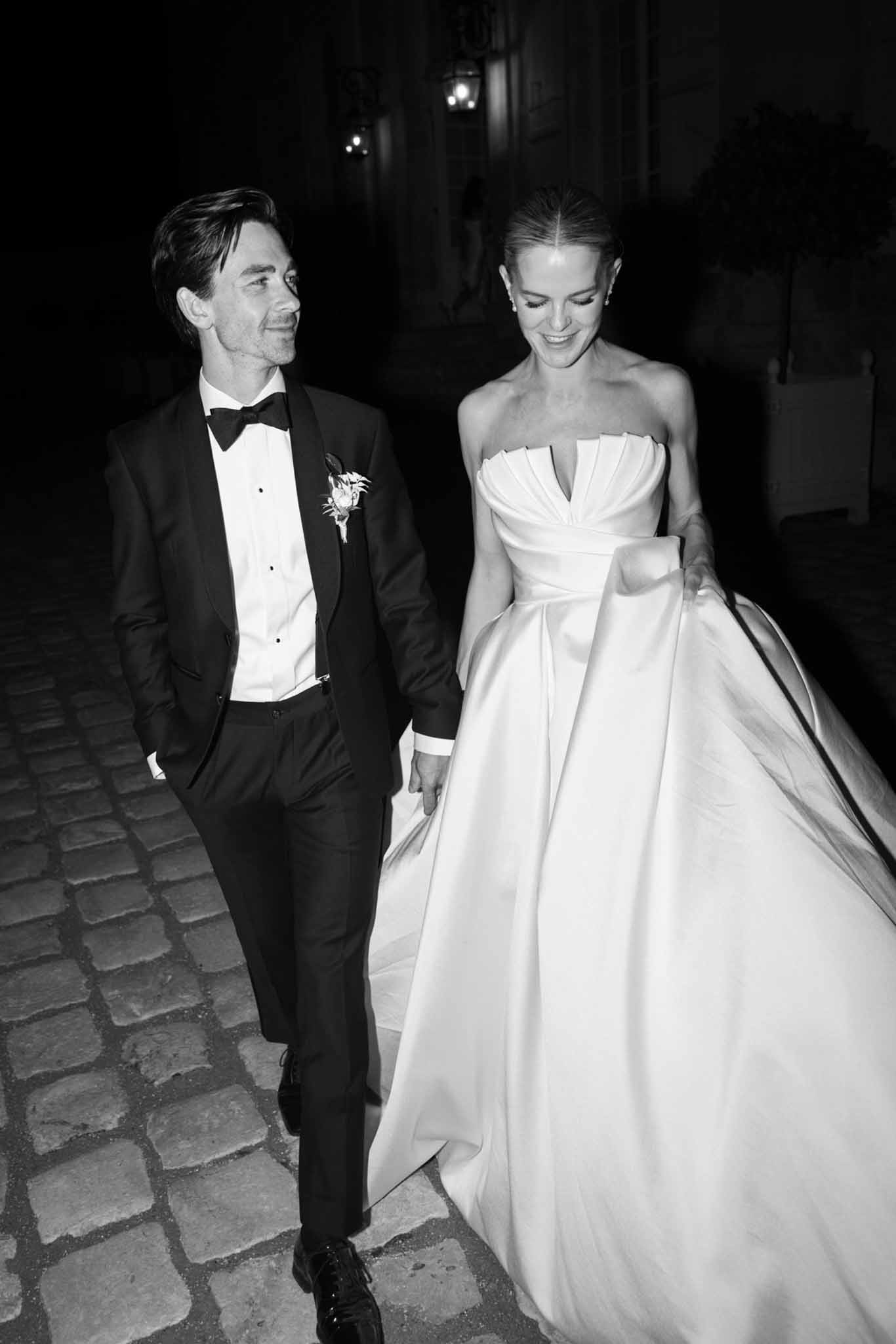 Bride and groom walking together down a cobblestone alleyway at night, lit by overhead lanterns, black and white photo