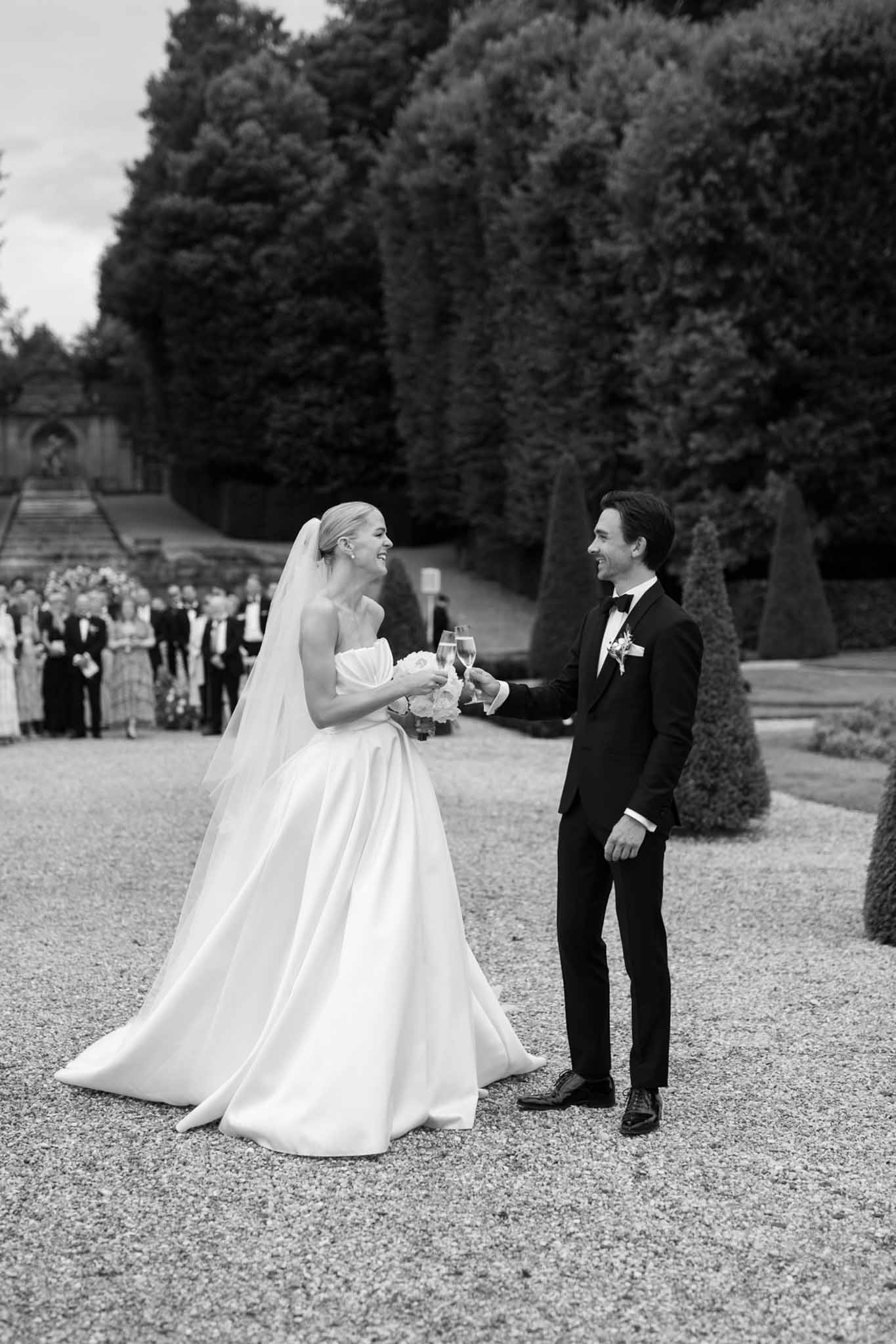 Bride and groom holding champagne glasses in formal garden, black and white, guests and cypress trees in background