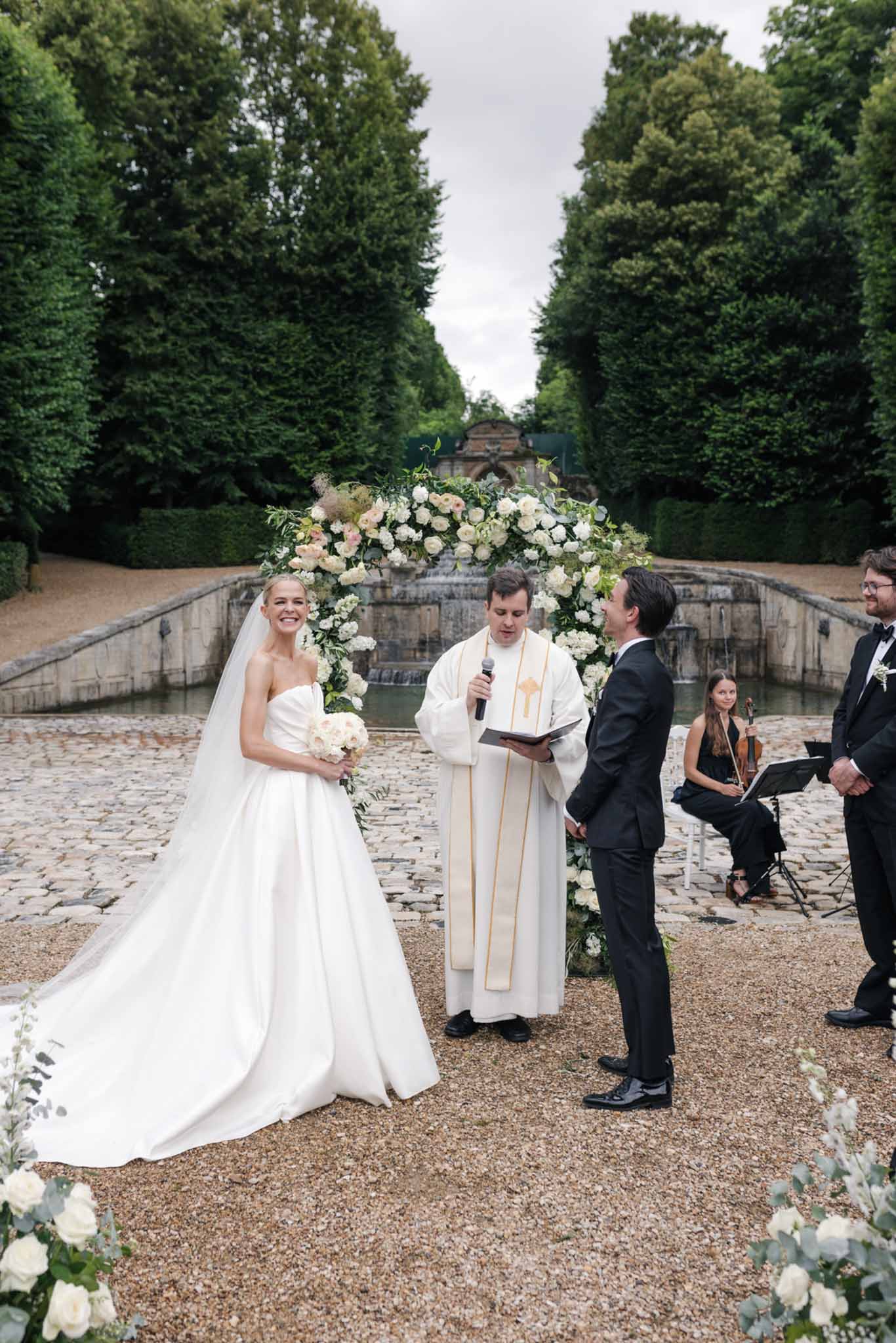 Bride and groom exchange vows in formal garden with rose-covered ceremony arch and manicured hedges