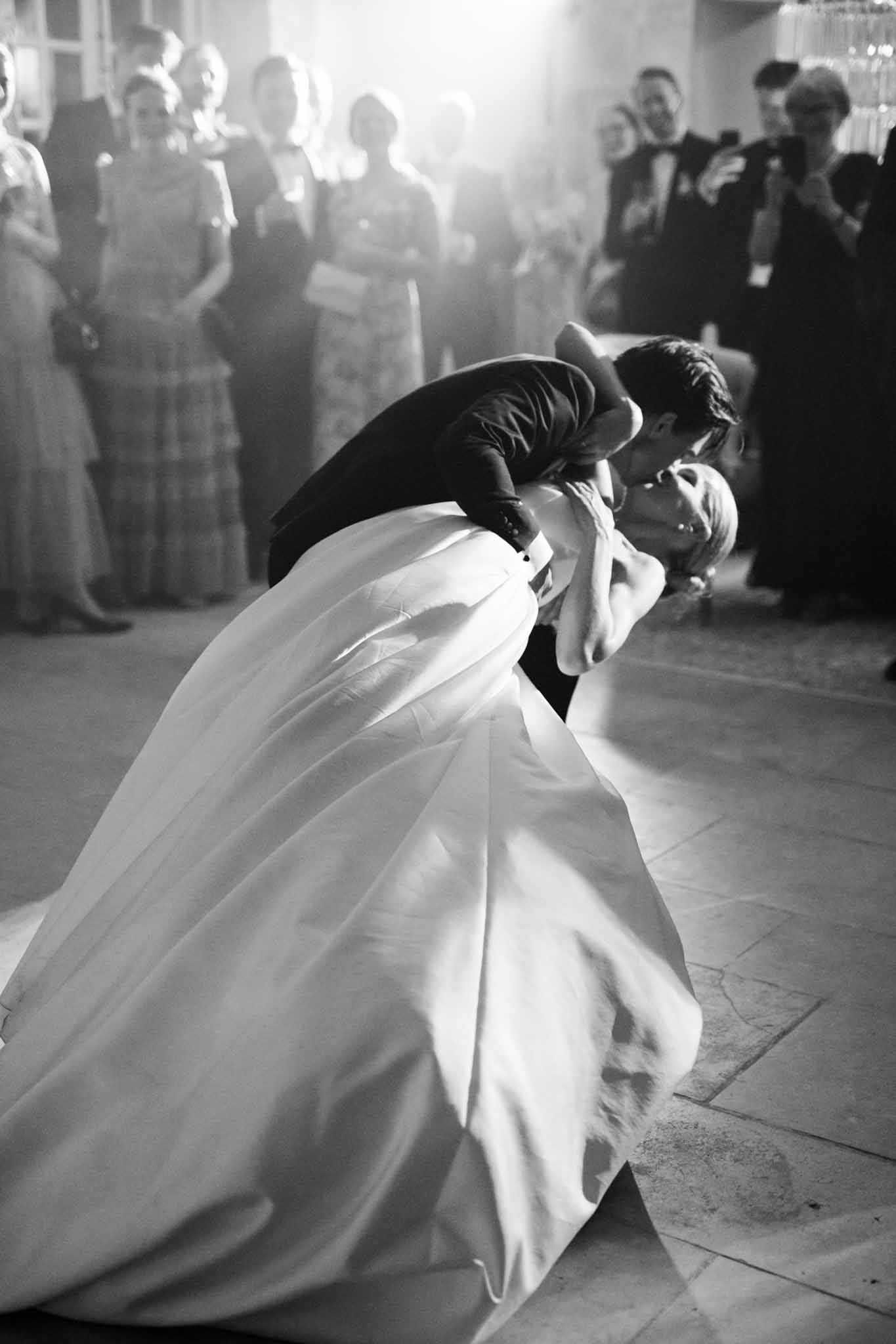 Black and white first dance: groom dips bride dramatically on an indoor dance floor while guests look on in a semi-circle.