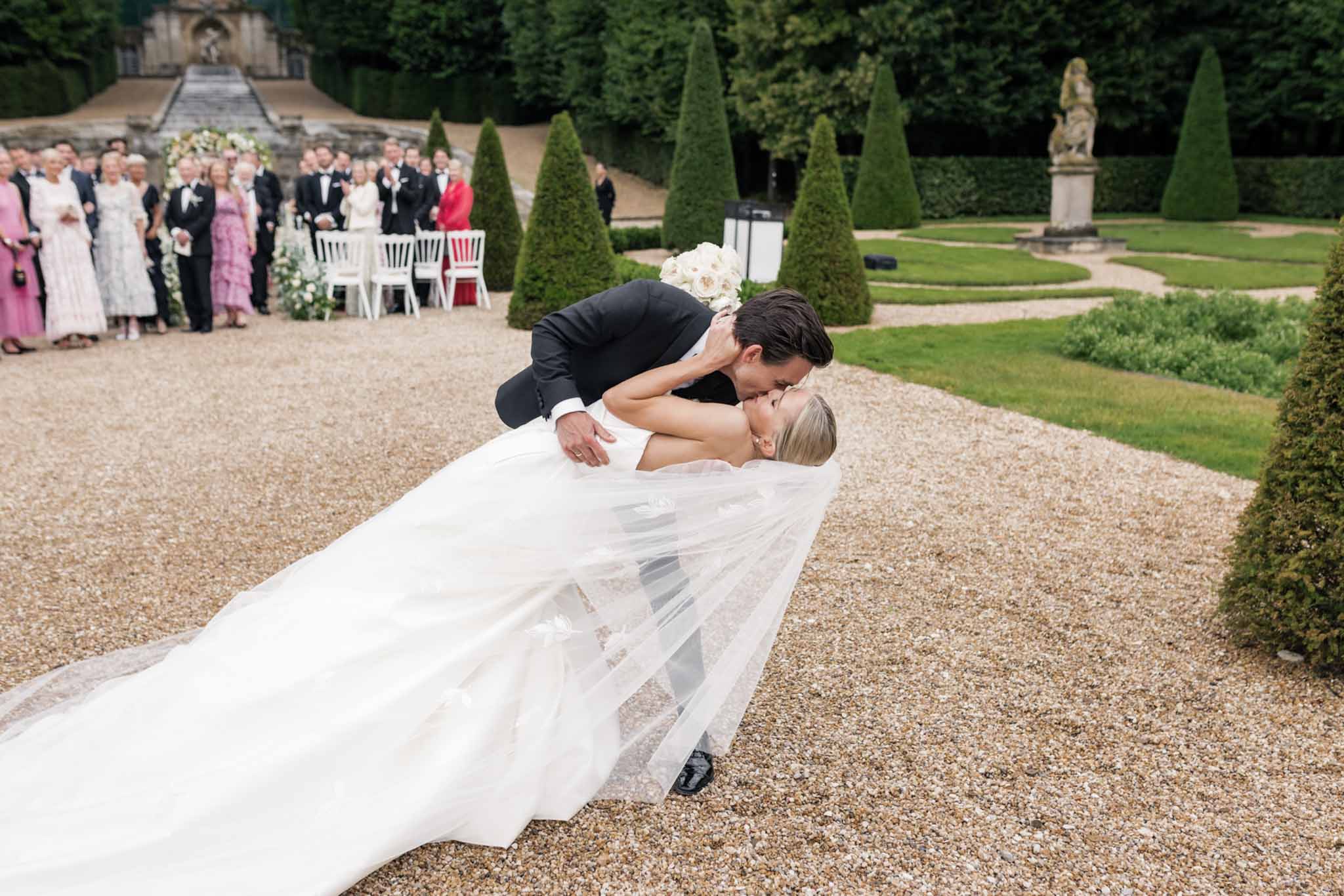 Groom dips bride on gravel courtyard as thirty guests watch, formal château garden with cone topiaries in background