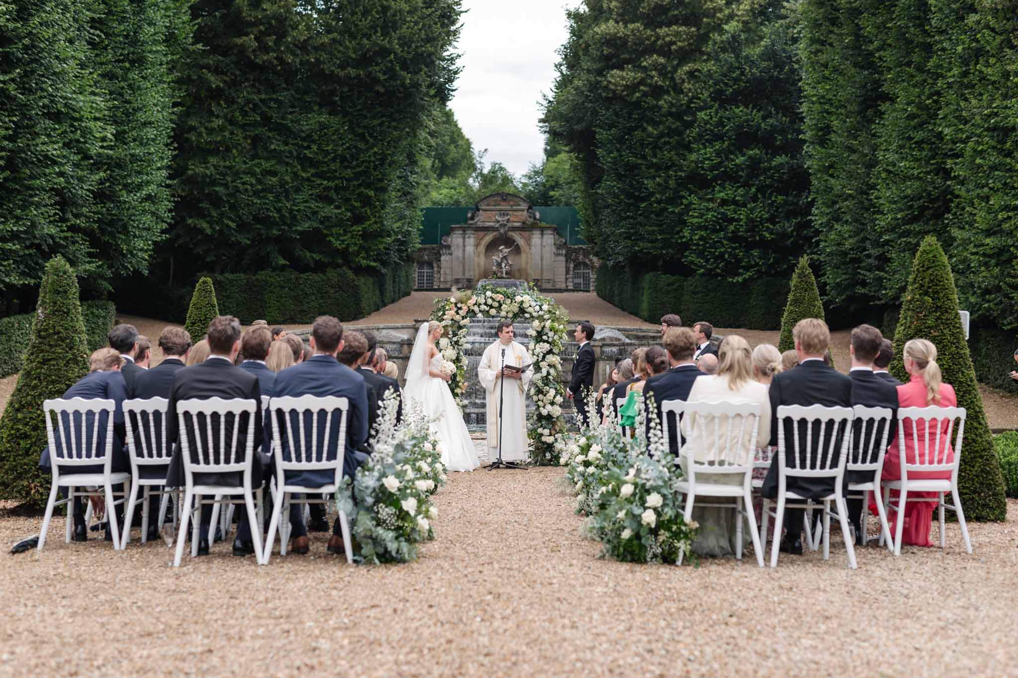 Outdoor ceremony in formal garden, couple at floral altar with 50 guests in white Chiavari chairs and clipped hedges