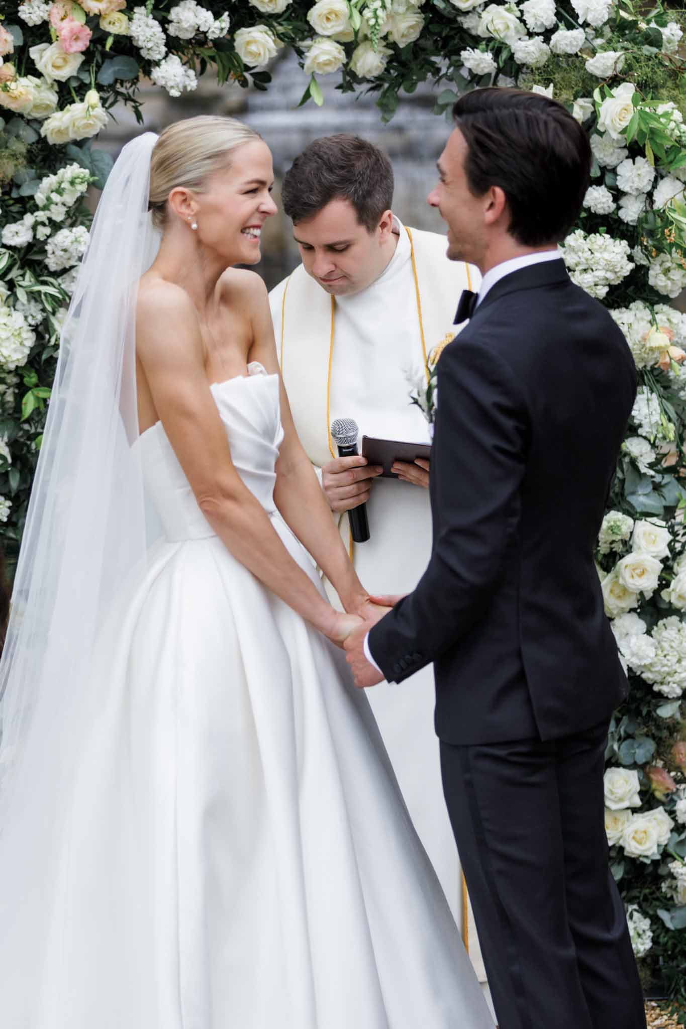 Bride and groom exchanging vows beneath a floral arch of ivory roses, white hydrangeas, and eucalyptus