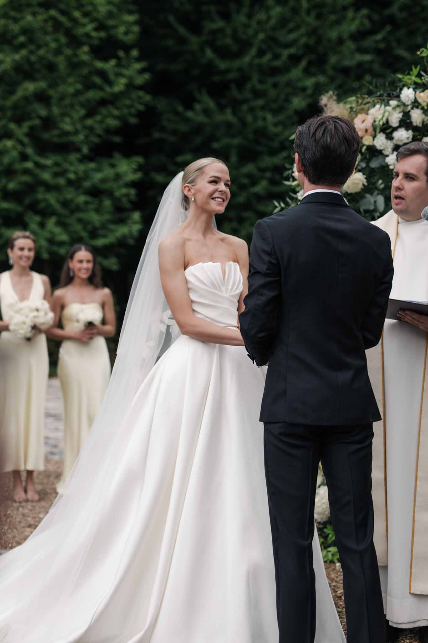 Bride and groom exchanging vows in formal garden ceremony with bridesmaids in cream dresses and white floral arch