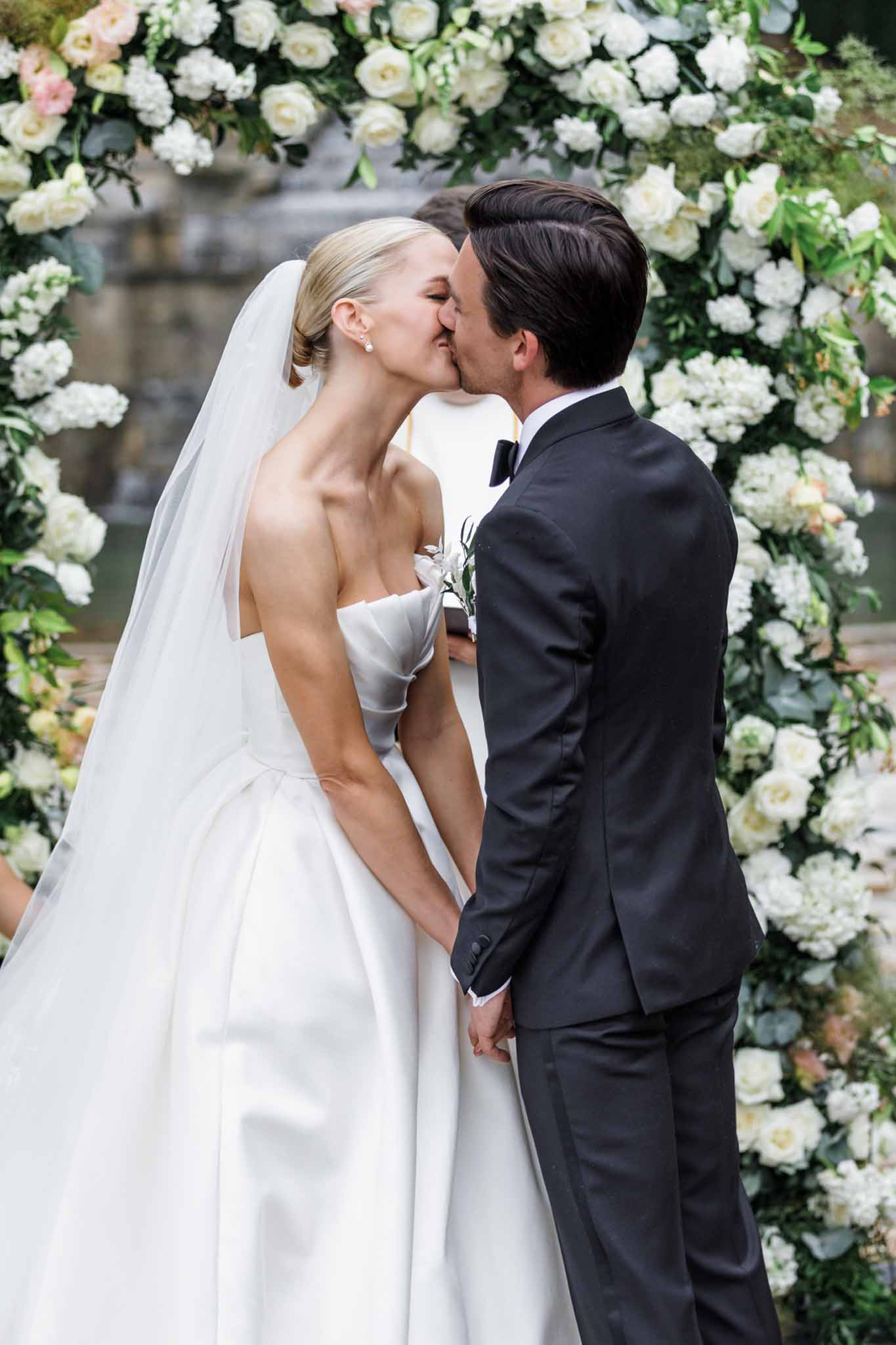 Bride and groom share first kiss under floral arch of ivory and blush roses at outdoor ceremony