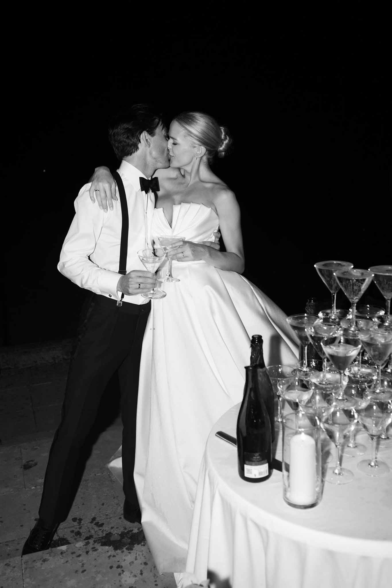 Black and white photo of bride and groom kissing while holding cocktails beside champagne glasses