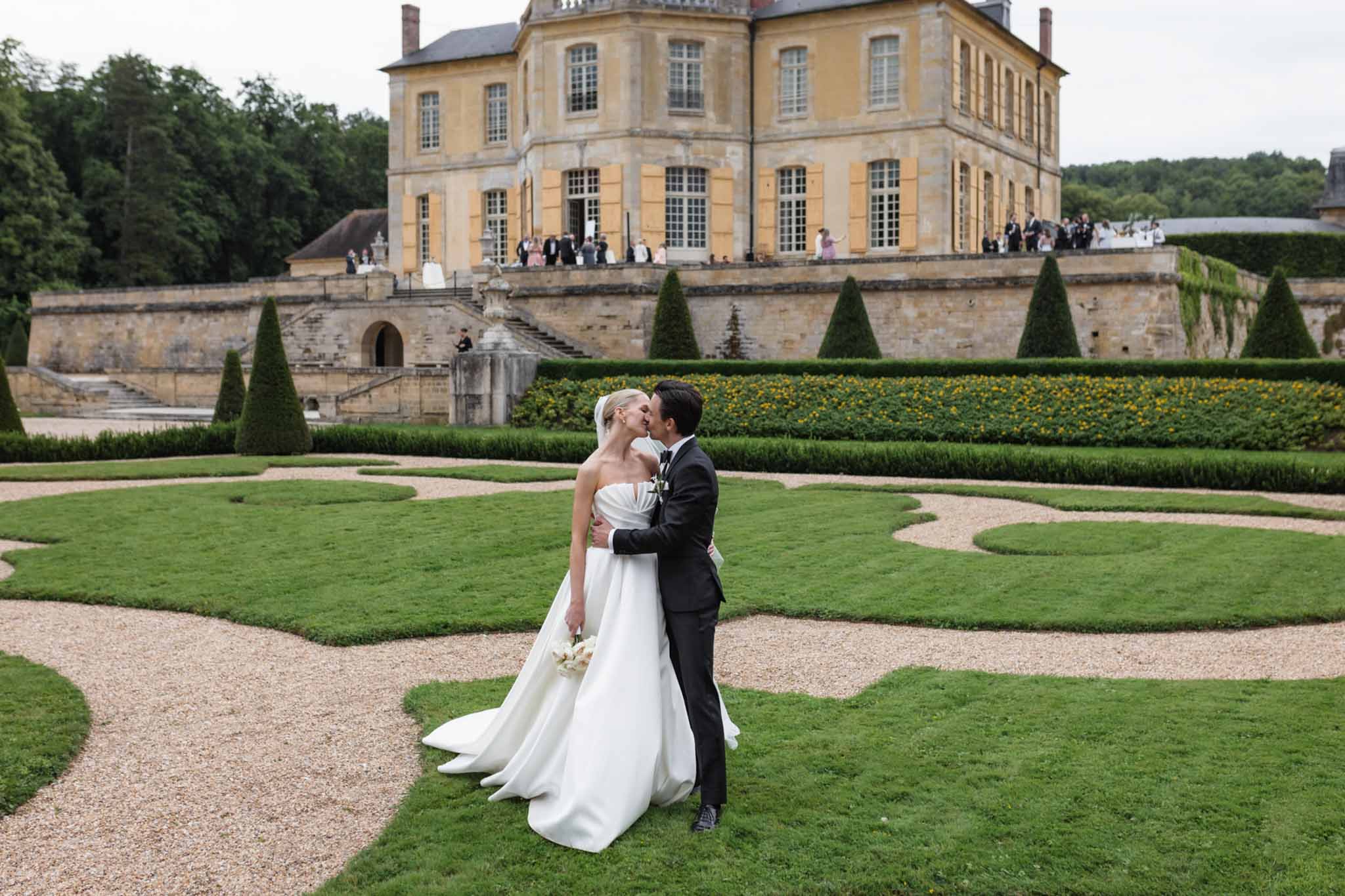 Bride and groom kissing in formal parterre garden, cream stone château with mansard roof and guests on terrace behind