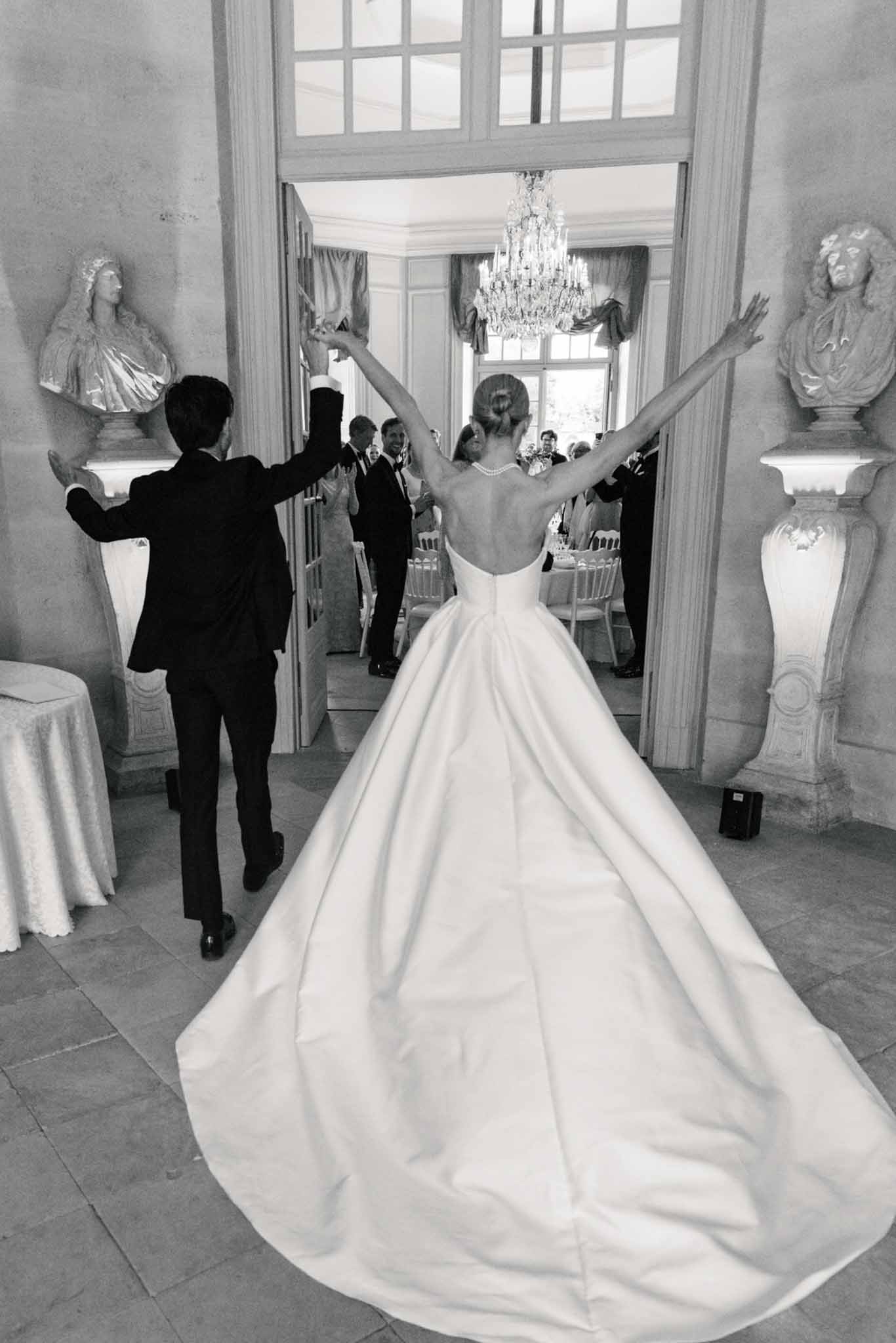 Black and white photo of couple celebrating with raised arms in neoclassical gallery with chandelier