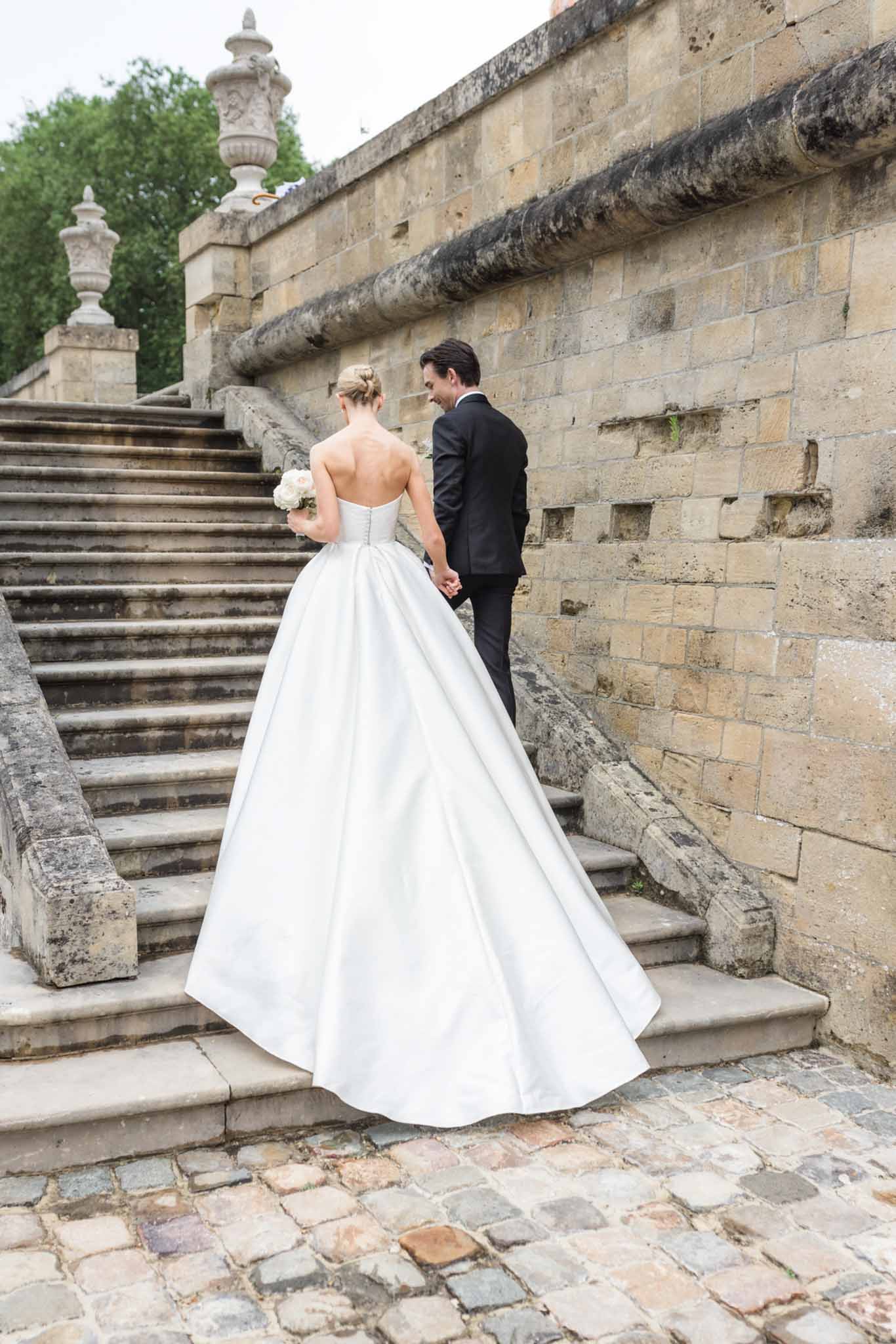 Bride in ivory ball gown and groom in black suit holding hands on stone staircase, photographed from behind