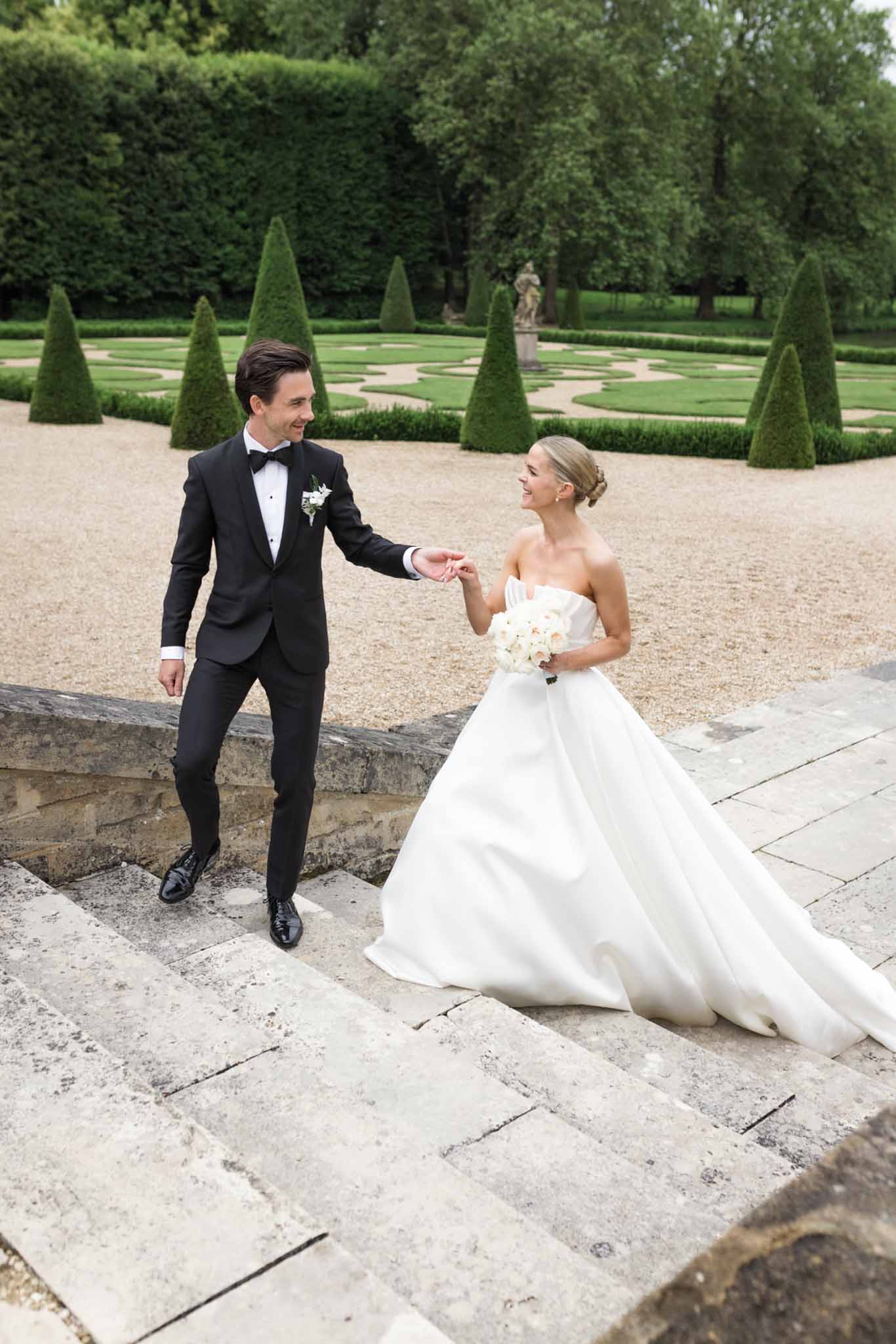 Bride in white ball gown and groom in black tuxedo walk gravel path with topiary cones and garden statue