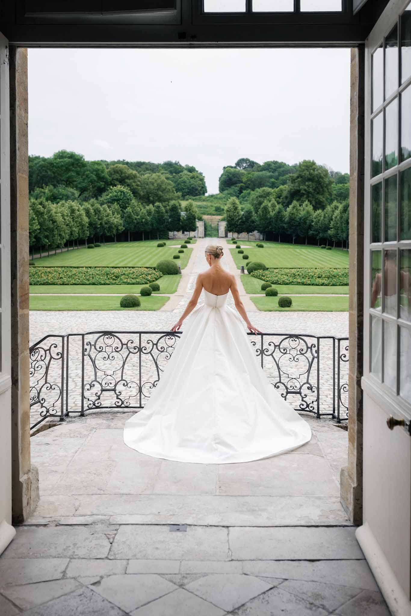 Bride in ivory ball gown on stone terrace gazing over formal French garden with topiary through open glass doors