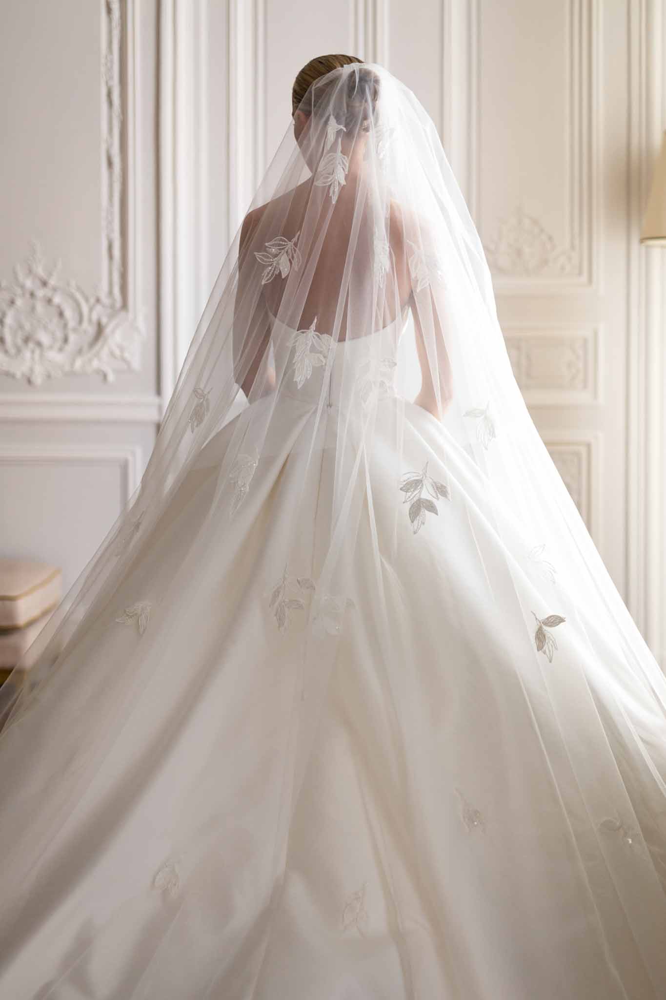 Rear-view portrait of bride in ivory ball gown with cathedral-length veil embroidered with gray-green leaf motifs in paneled white interior