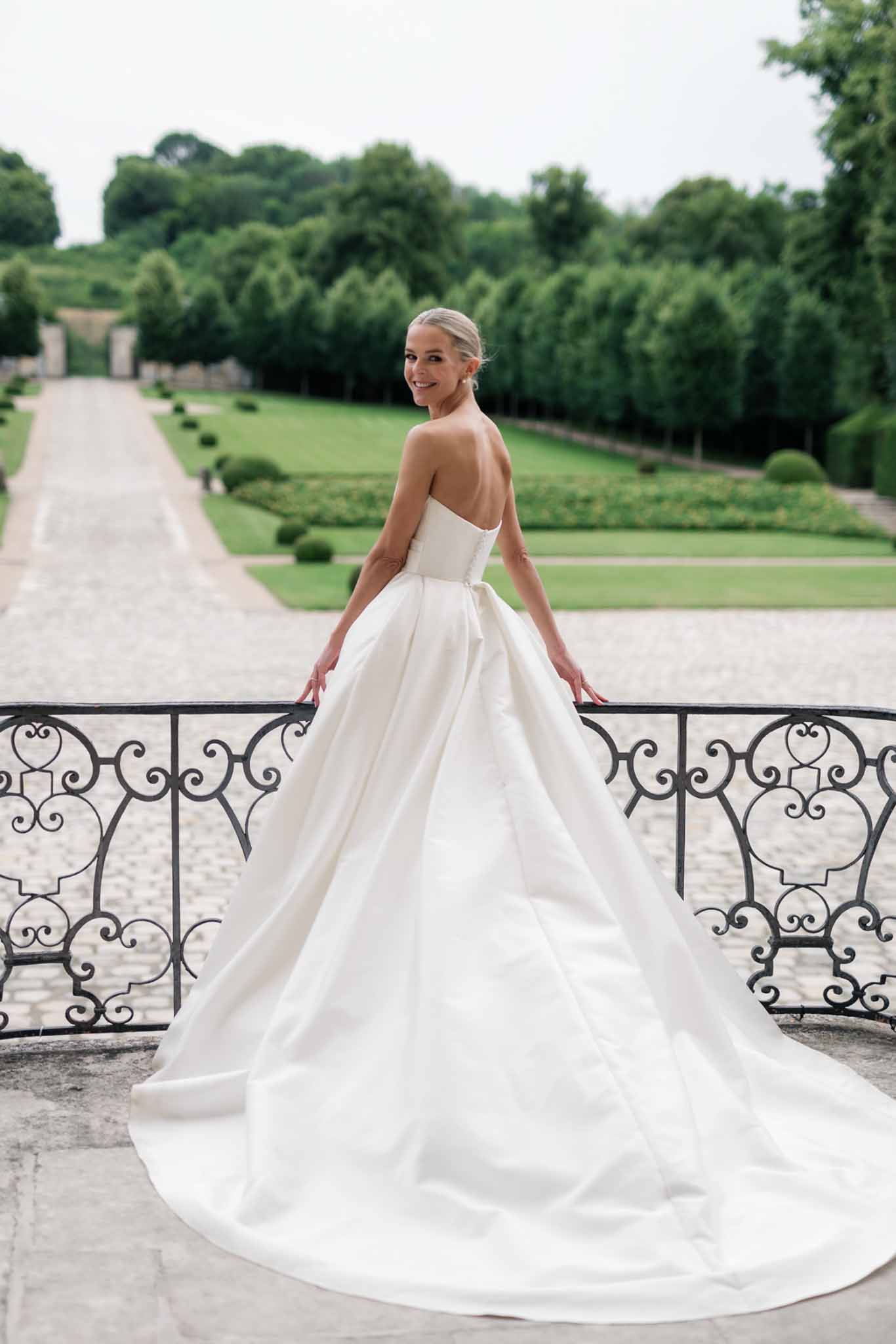 Bride in ivory ball gown at ornate wrought-iron railing on stone terrace overlooking formal gardens and château beyond