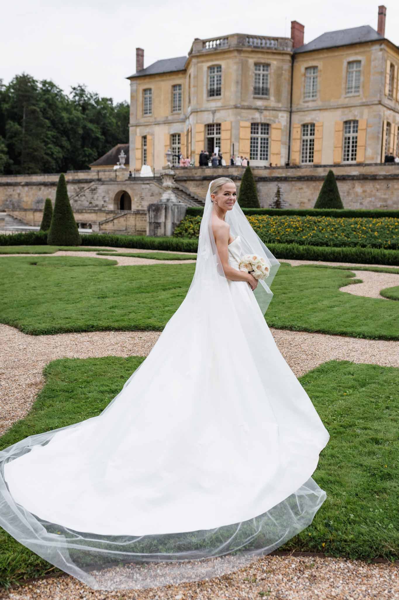 Bride in long white gown and full-length veil standing in garden at Château de Villette, Paris