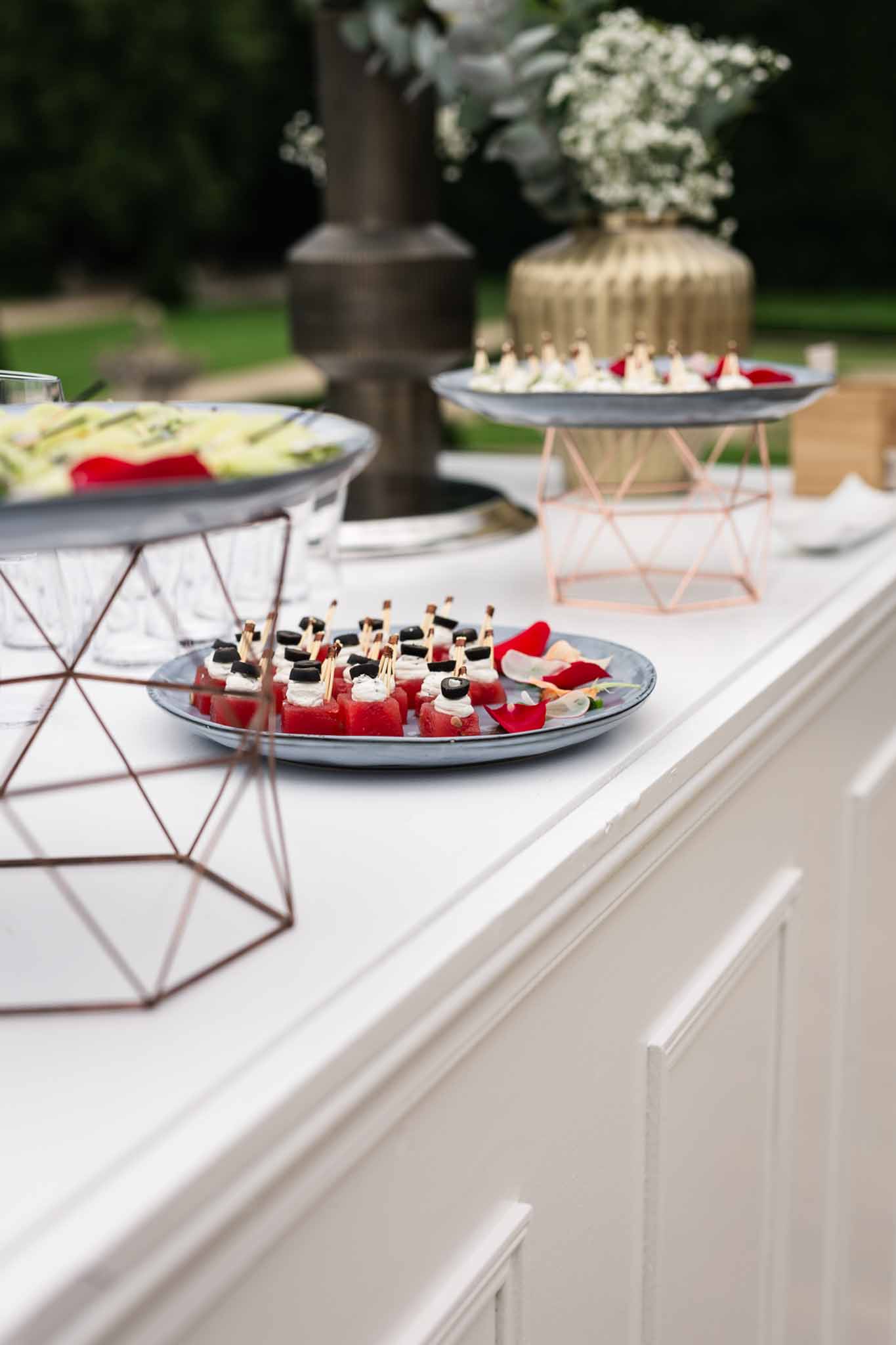 Cocktail hour display with canapes on slate plates and rose gold geometric stands, white floral urns behind on terrace