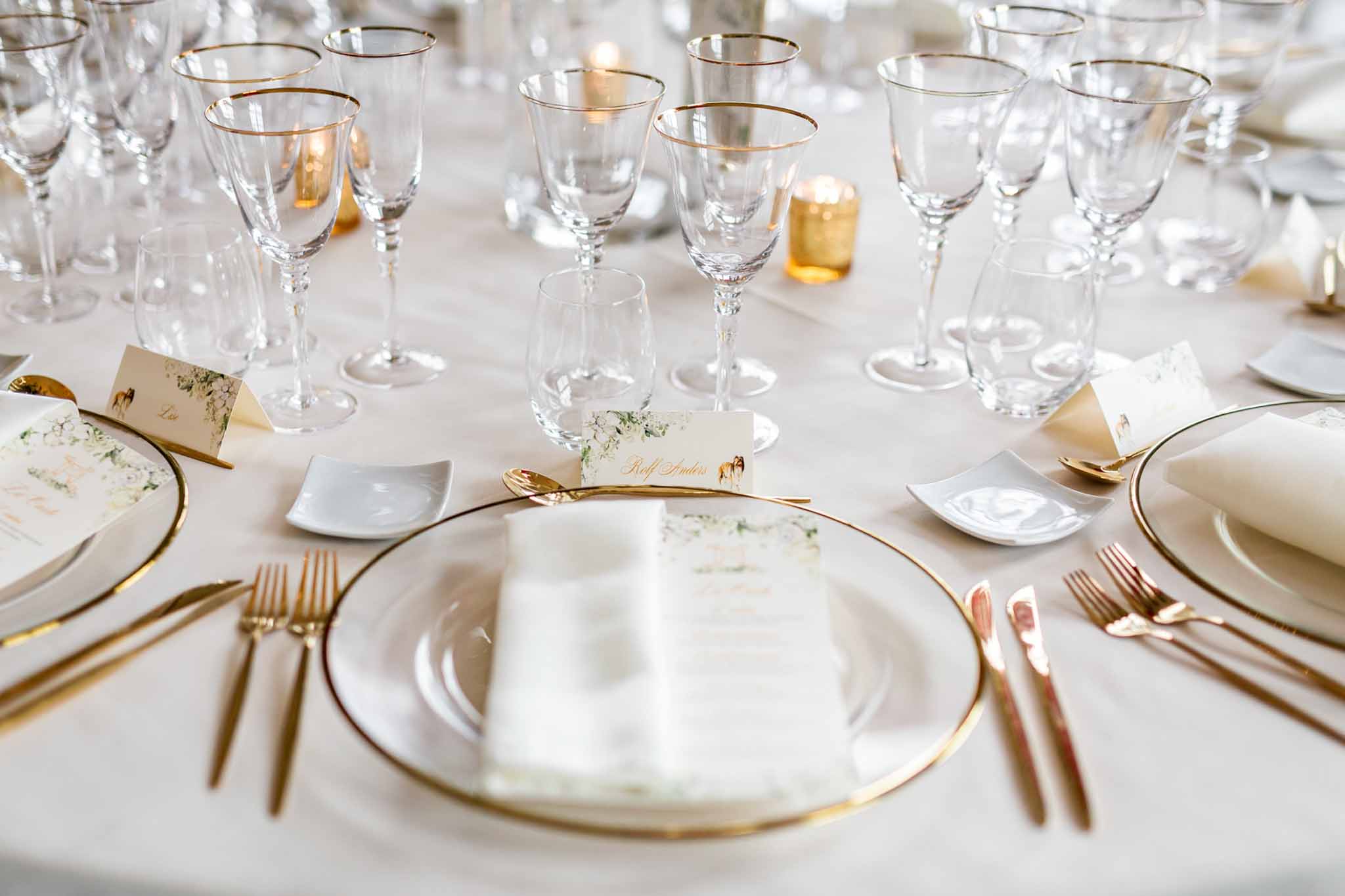 Close-up of reception table setting with gold-rimmed chargers, gold flatware, crystal glasses, and white place cards