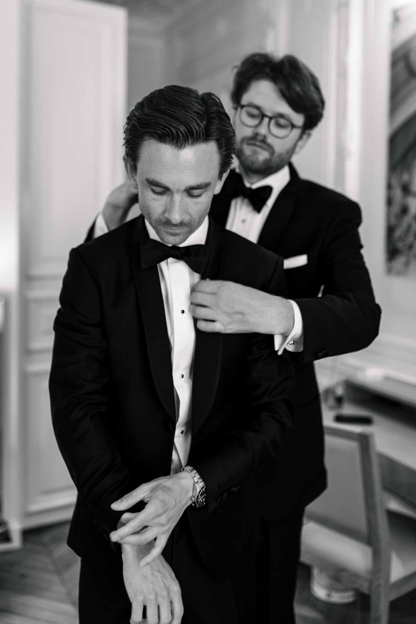 Black and white photo of groomsman adjusting groom's black tuxedo jacket in hallway before ceremony