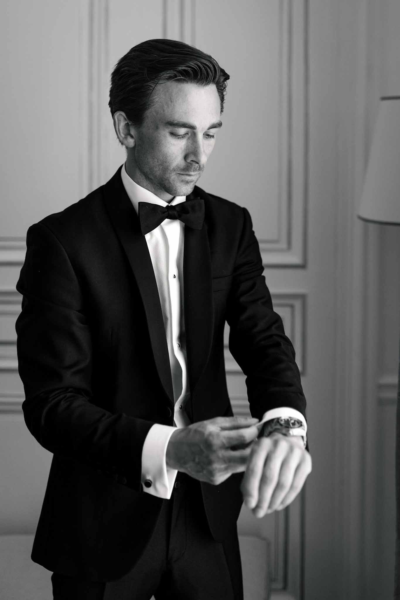 Groom in black tuxedo checking his wristwatch, black and white portrait in panelled room