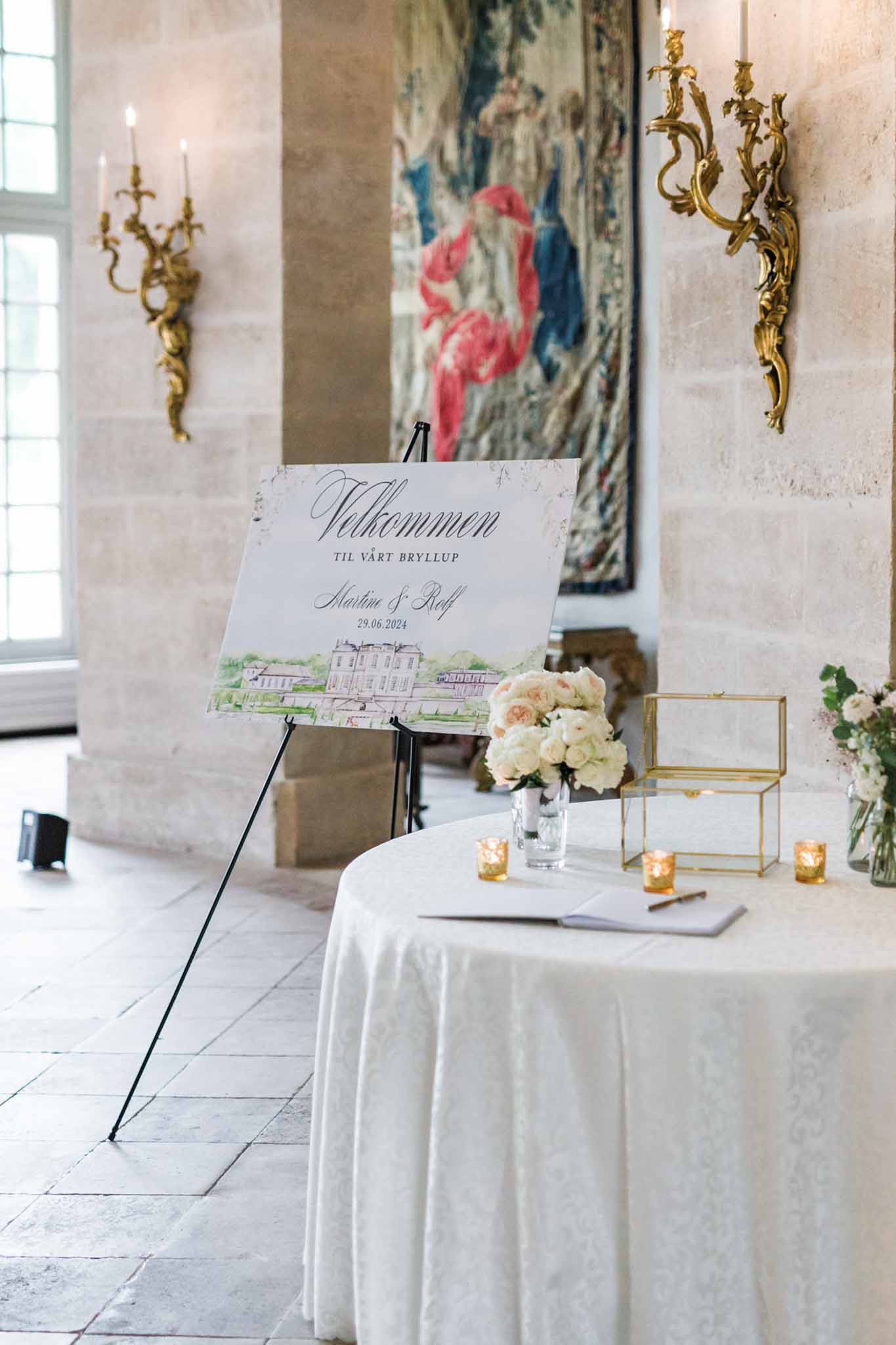 Wedding welcome display with calligraphy sign on easel, cream roses, and guest book in classical venue interior