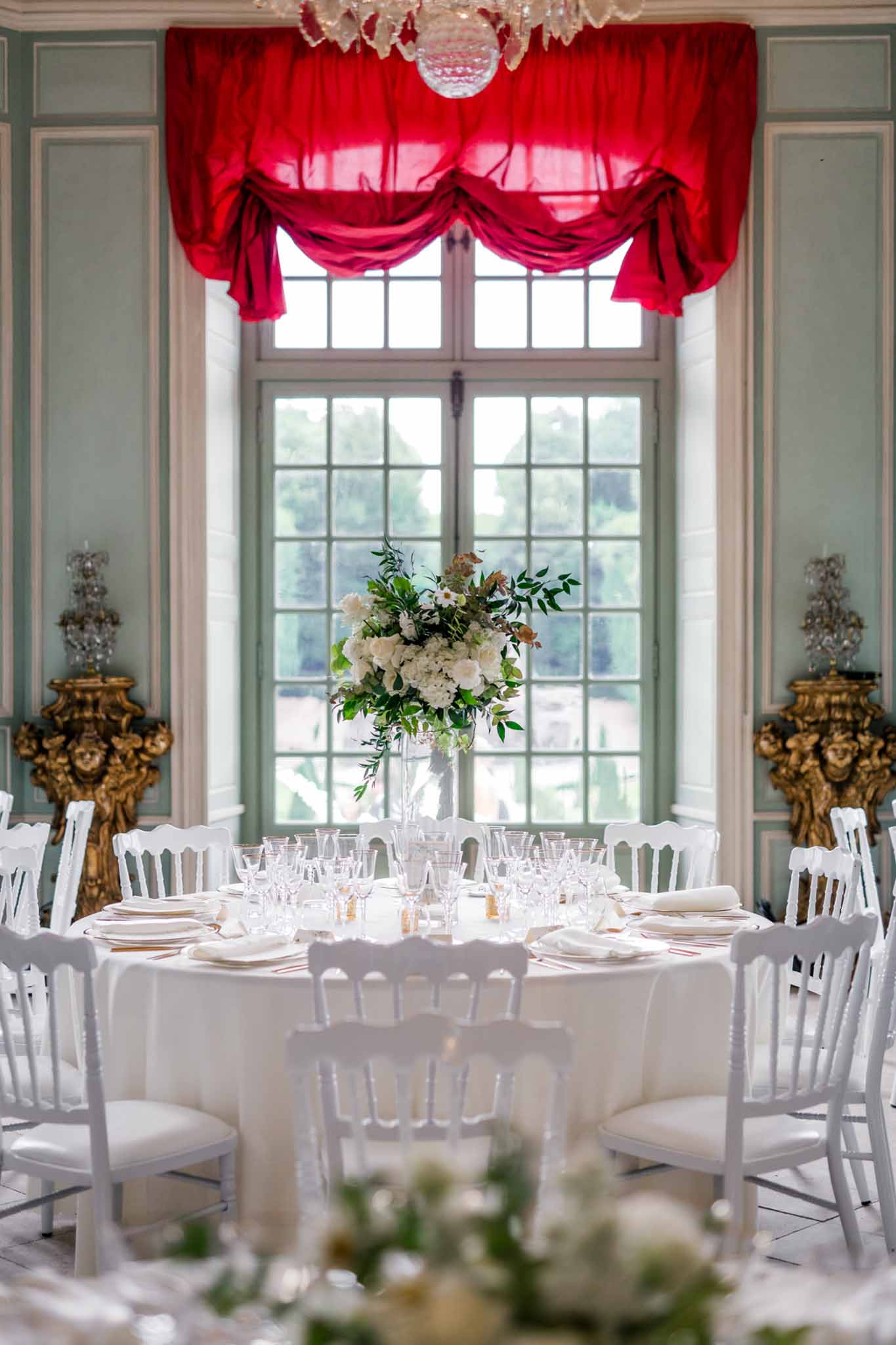 Round reception table with ivory linens and Chiavari chairs beneath a crystal chandelier and red velvet curtains