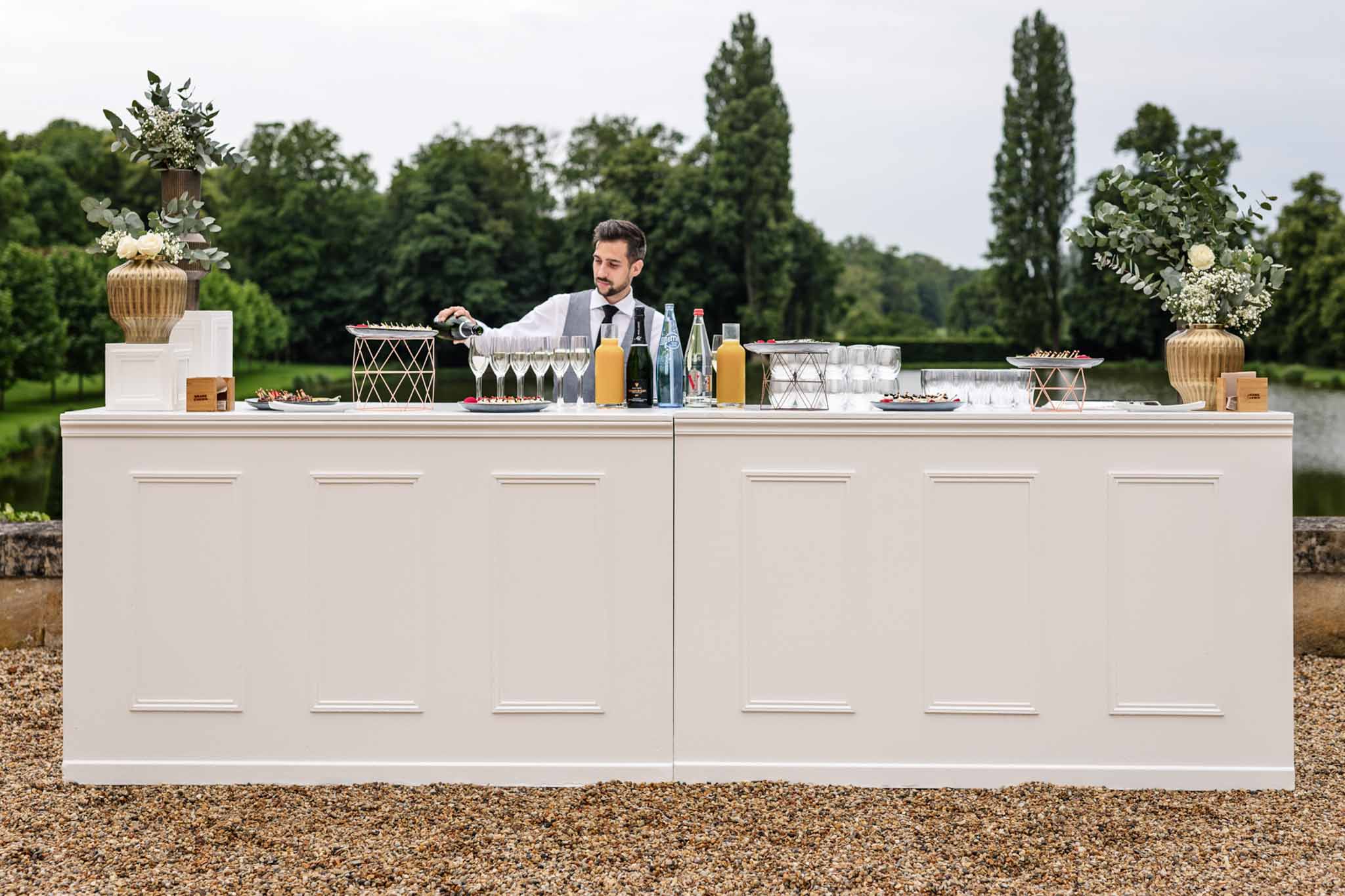 Bartender at cream outdoor bar with champagne, cream rose arrangements in gold urns, and riverside poplar trees