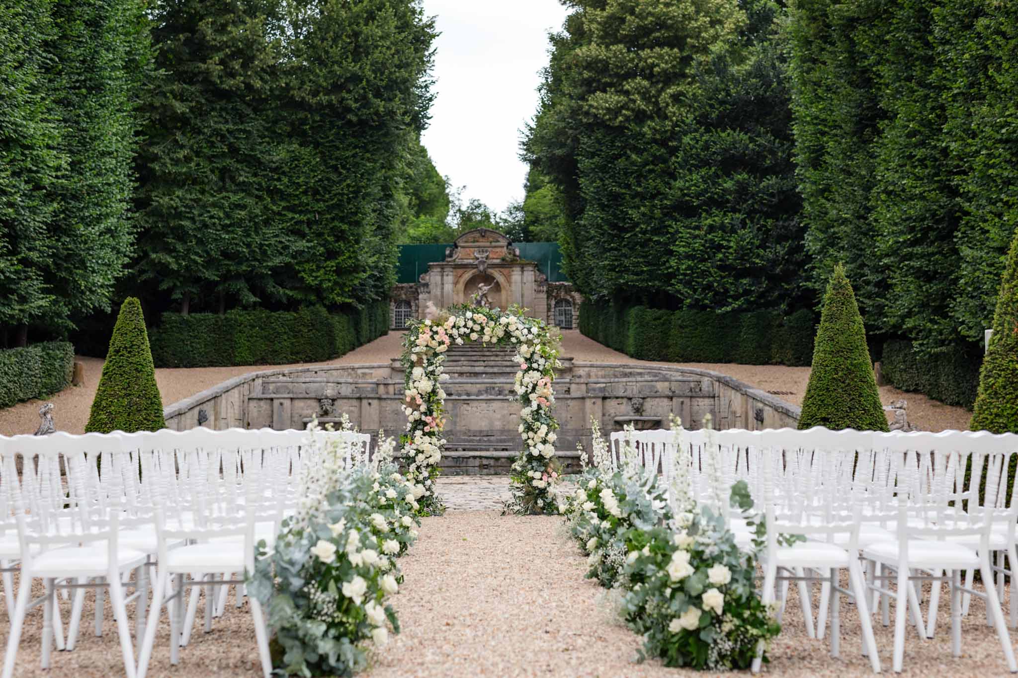 French garden ceremony with white Chiavari chairs, ivory and blush floral arbor and classical stone fountain