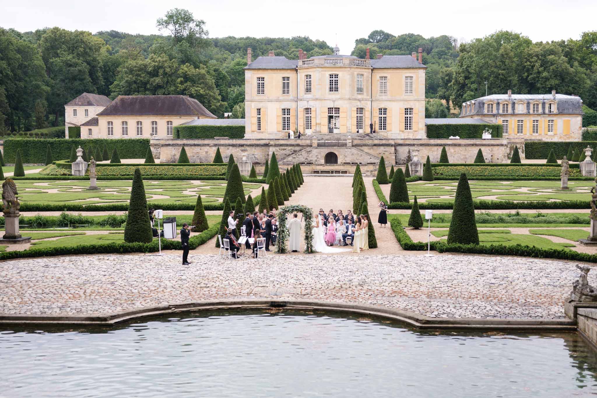 Wedding ceremony in chateau garden viewed across reflecting pool with greenery arch