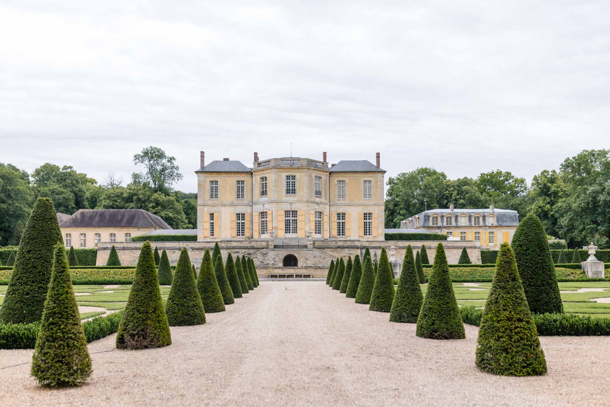 Classical French château with butter-yellow facade and mansard roof viewed from formal garden with cone topiaries