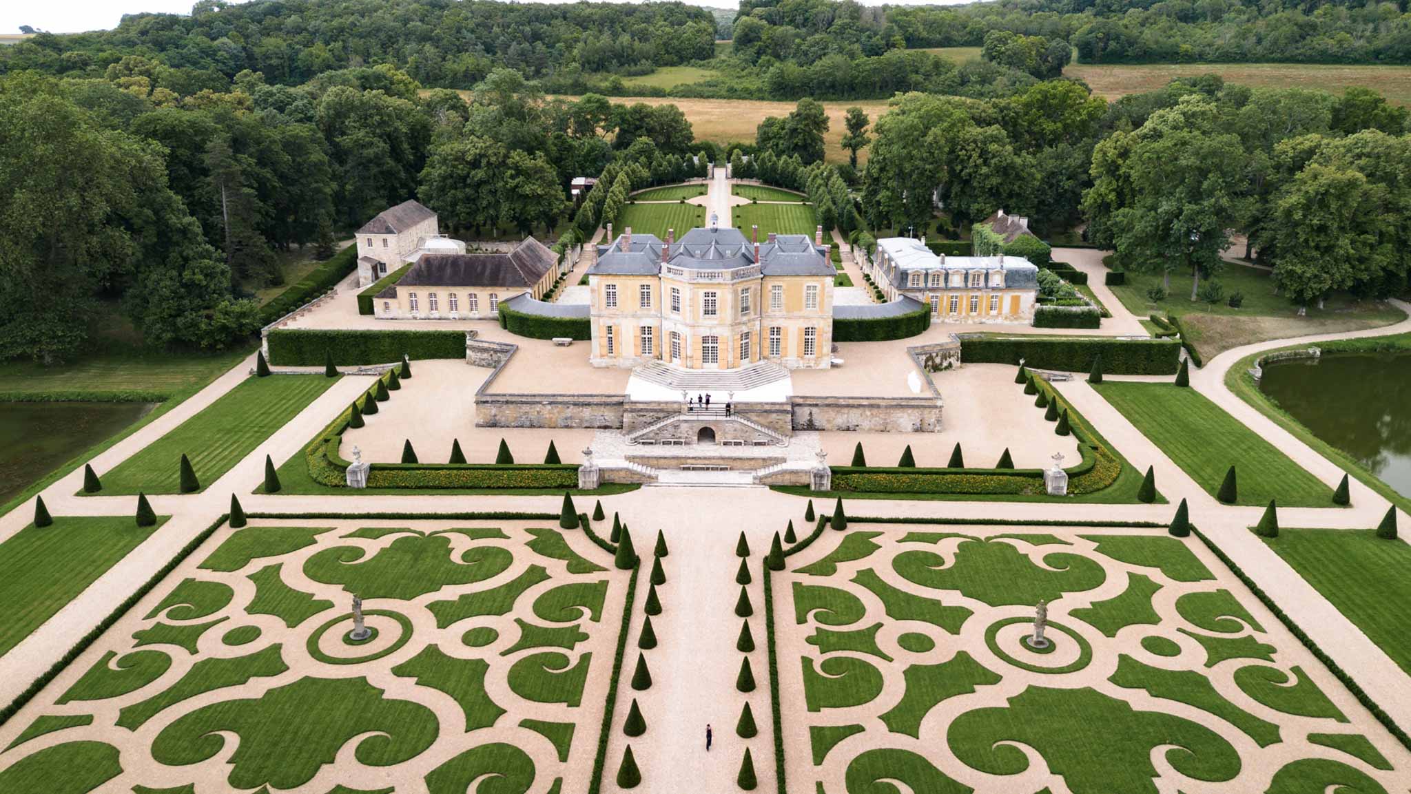 Aerial view of grand French chateau with formal baroque parterres, topiary cones, cross-shaped water feature, and woodland