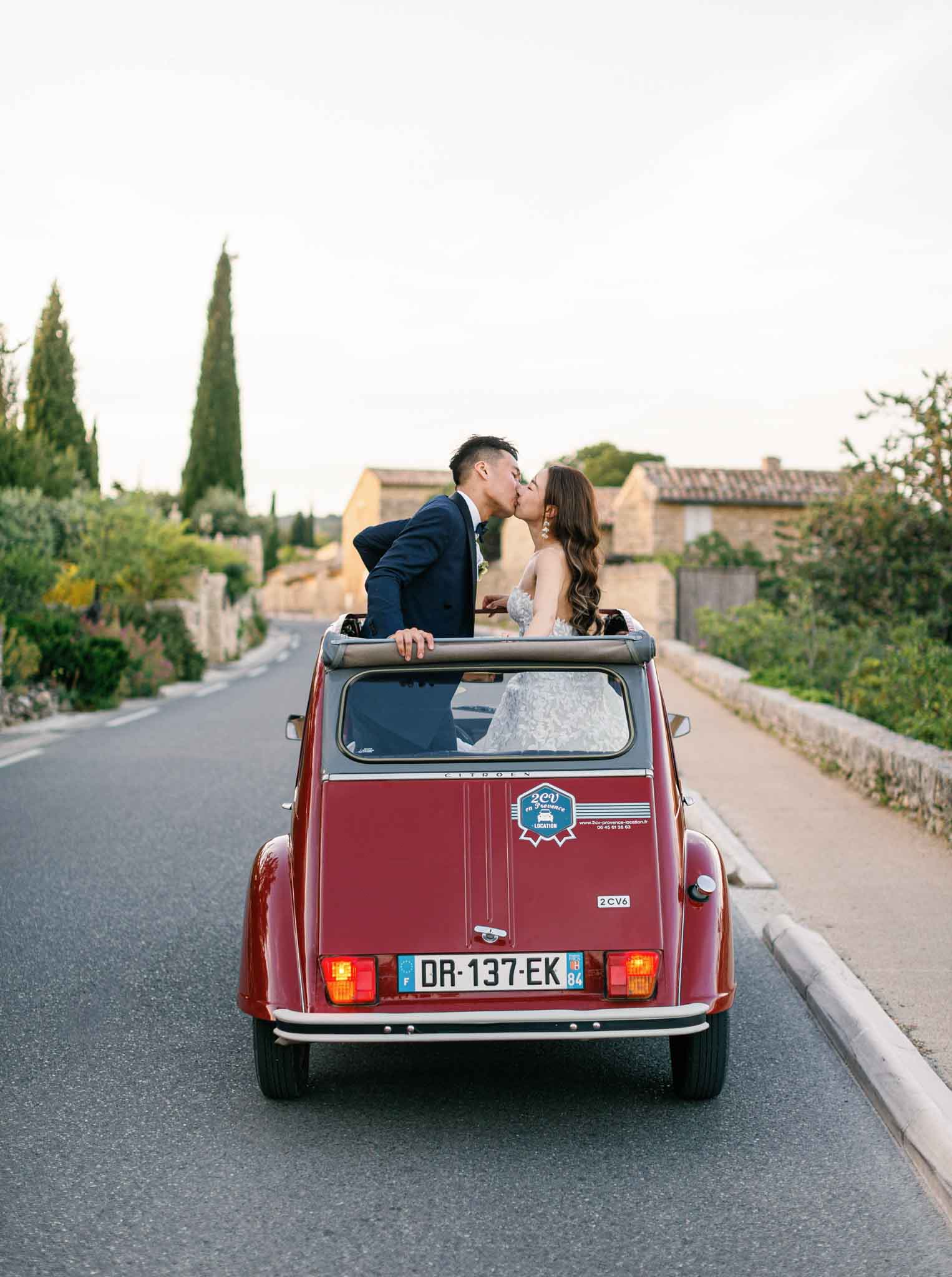 Couple kissing through sunroof of burgundy vintage Citroen 2CV on Provencal road lined with cypress trees