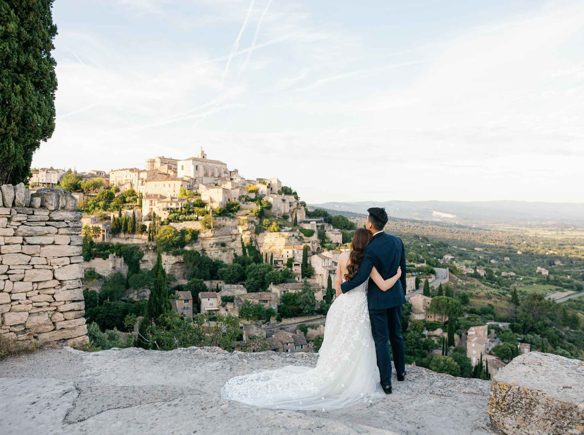 Bride in ivory lace gown and groom in navy suit on rocky hilltop overlooking honey-colored Provencal village