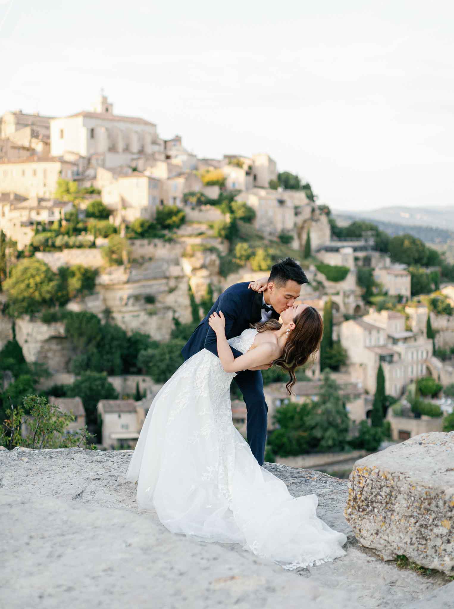 Groom dipping bride on stone overlook with medieval Provencal hilltop village and bell tower in background