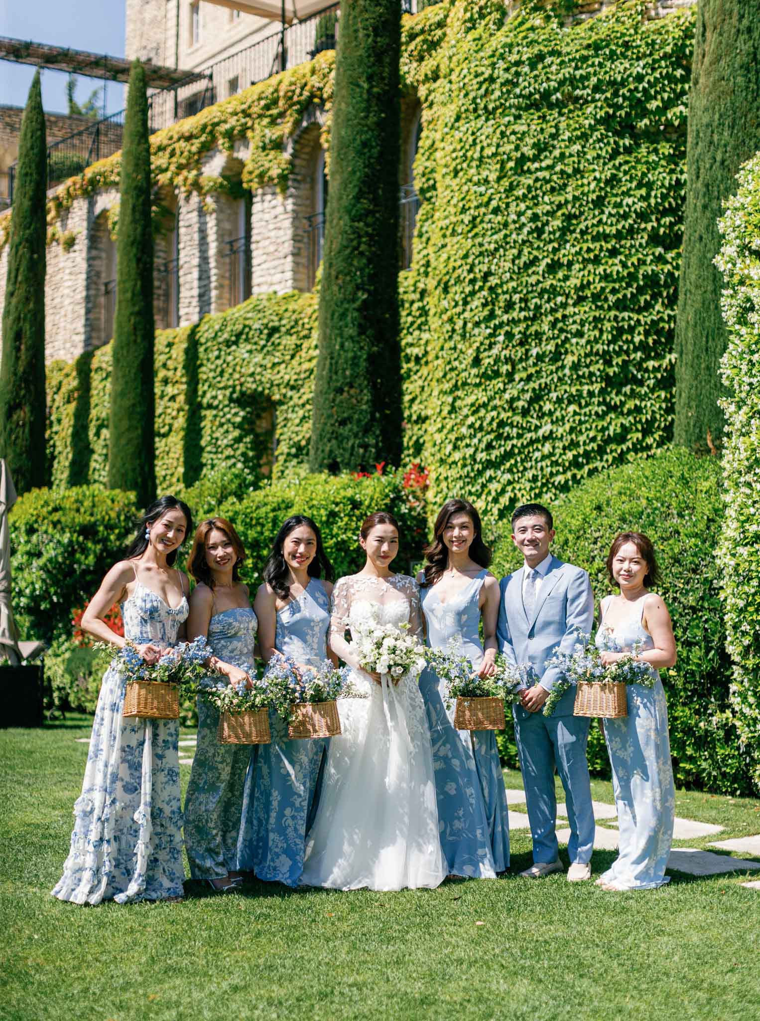 Bride and seven attendants in blue floral dresses stand in a line on a manicured lawn with ivy-covered stone building