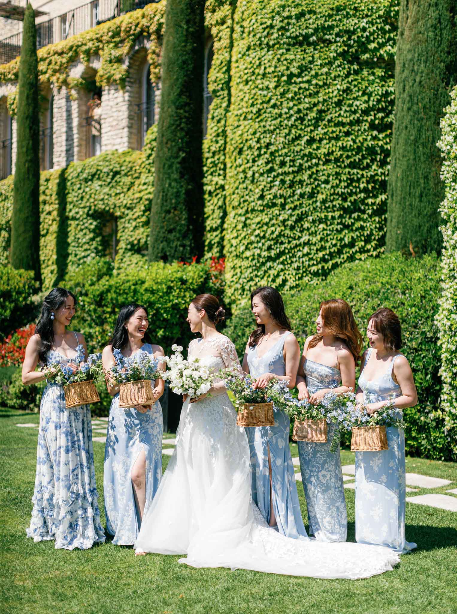 Bride in ivory lace gown with five bridesmaids in dusty blue floral dresses posing in a manicured garden with ivy-covered walls