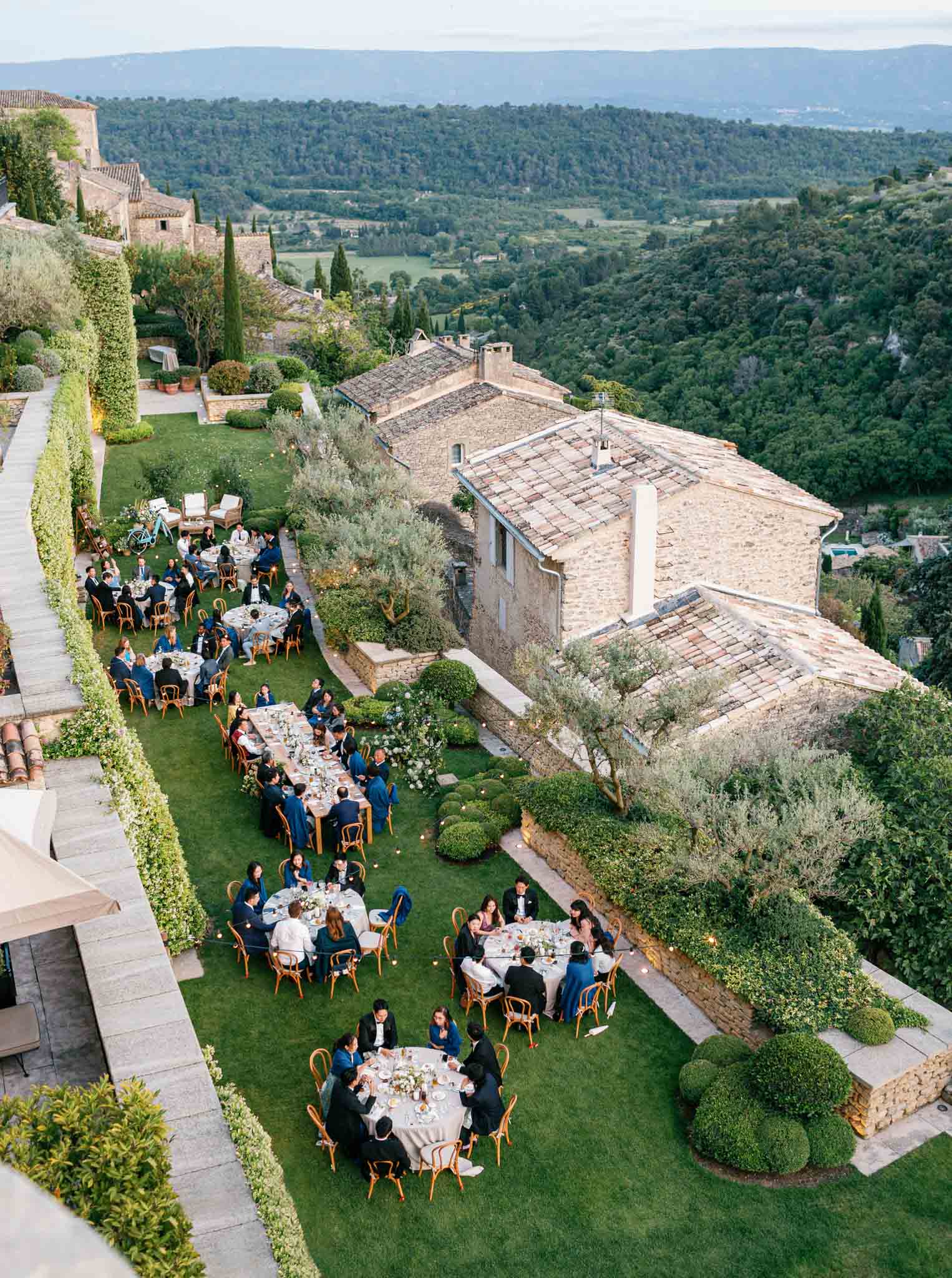 Aerial view of garden reception with round tables and gold rattan chairs at stone villa overlooking forested valley
