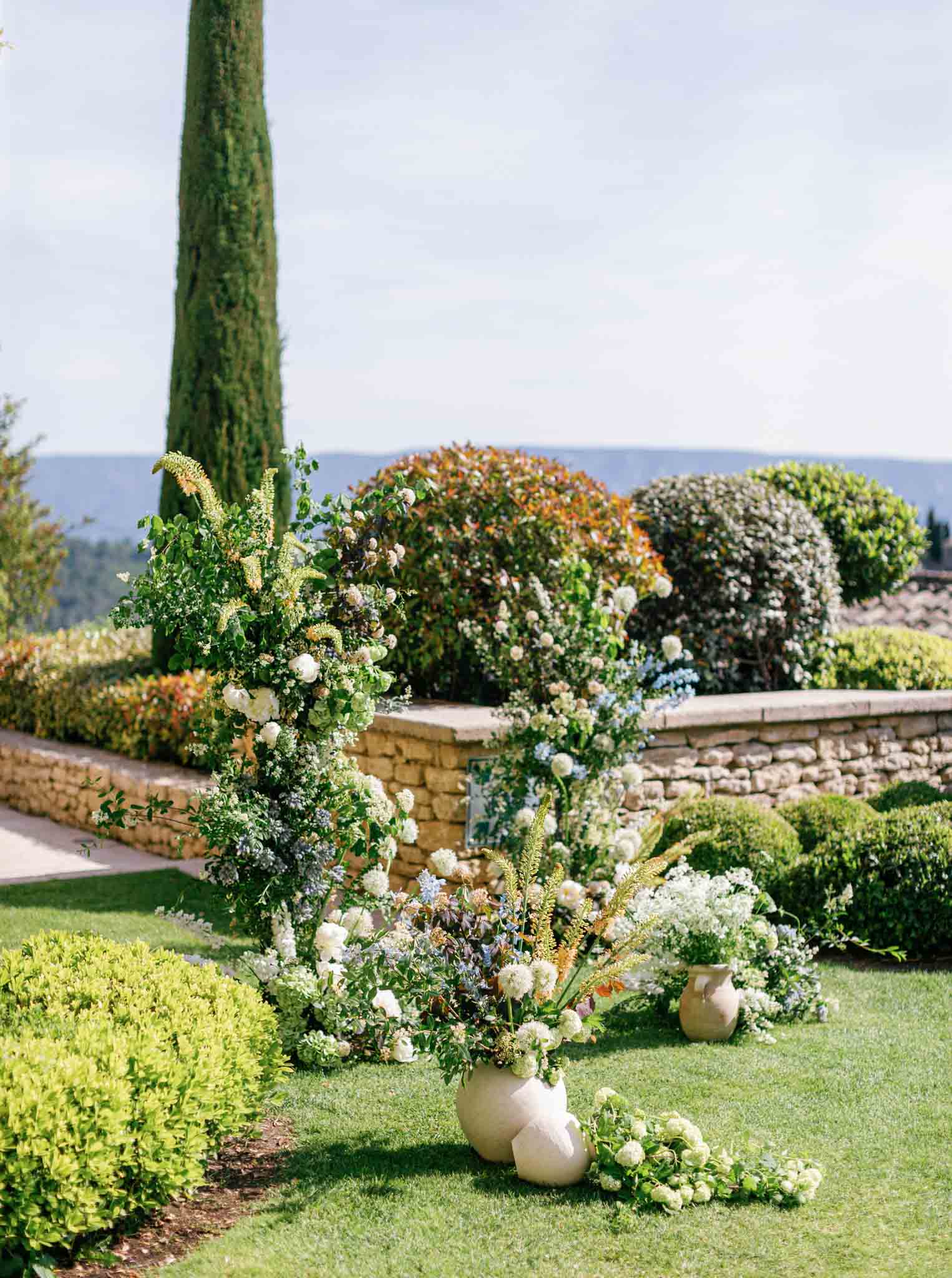 Formal Mediterranean garden with stone urns, cypress tree, boxwood topiaries, and water view at Gordes