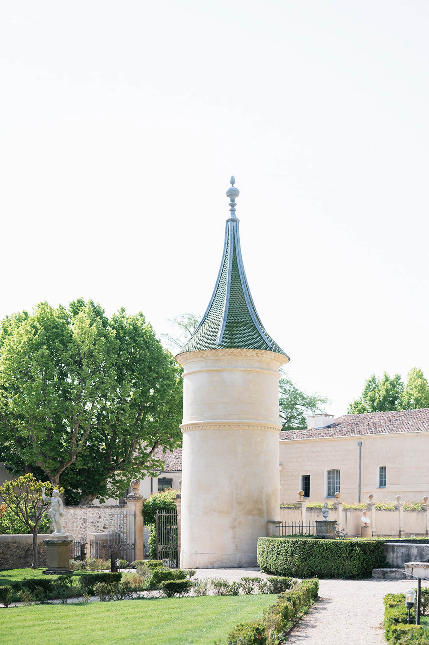 French chateau stone turret with pointed green roof flanked by formal boxwood gardens and gravel path